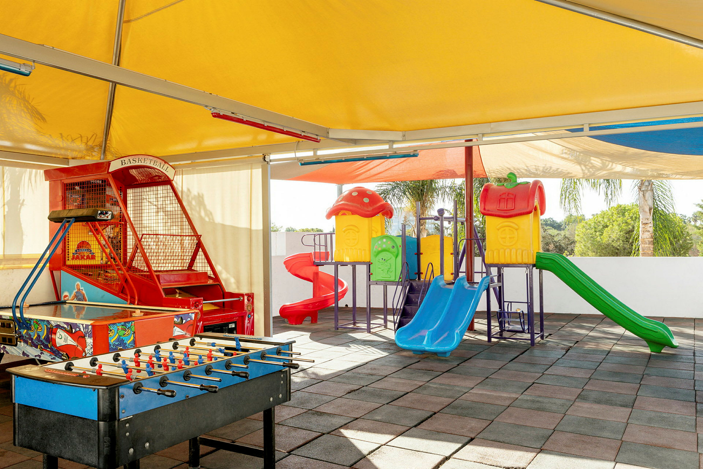 Billede af en udendørs legeplads med legehuse, rutsjebaner, basketballspil og et bordfodboldspil. (AI-genereret tekst)
