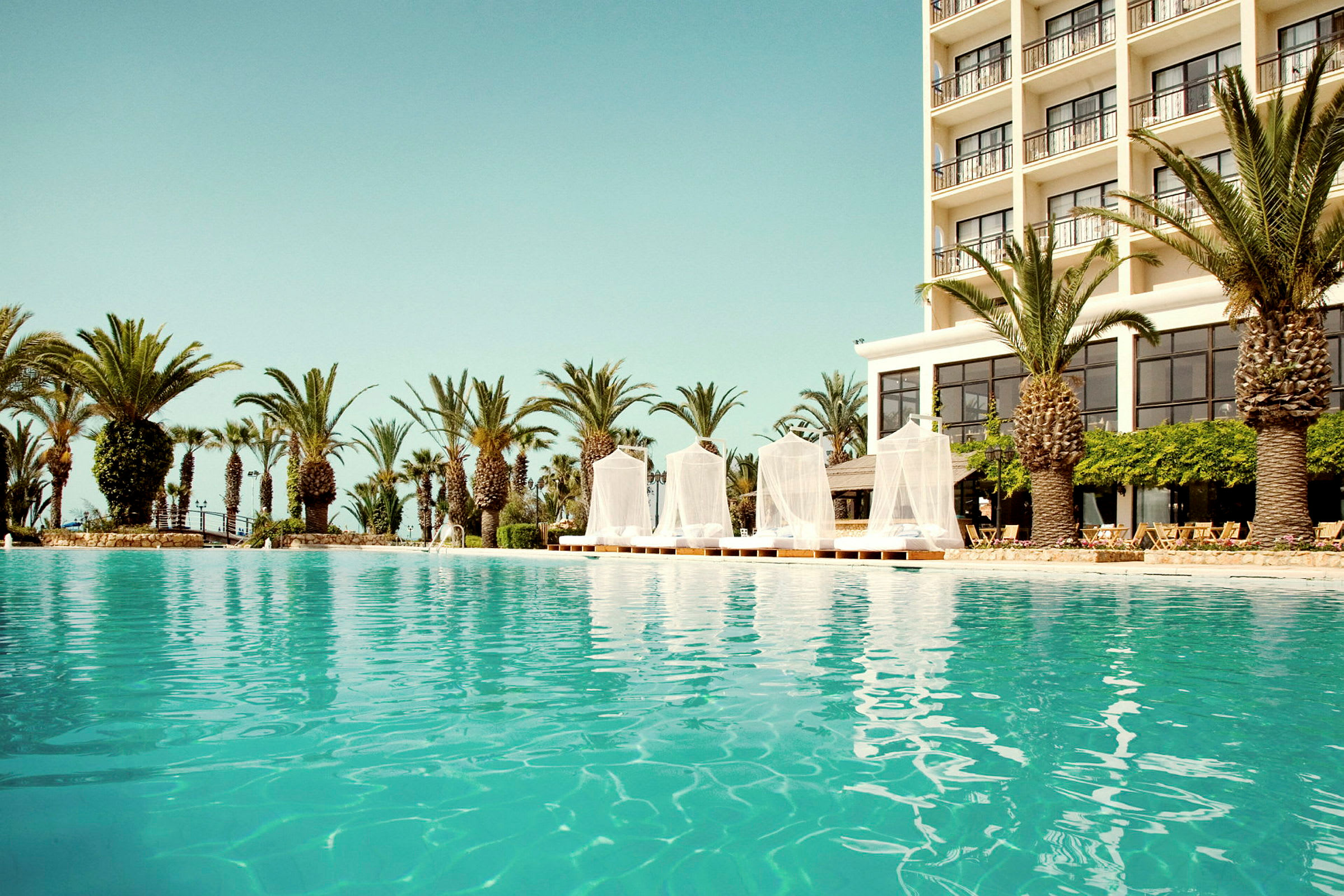 Udendørs pool med et hotel i baggrunden og omgivende palmetræer. (AI-genereret tekst)