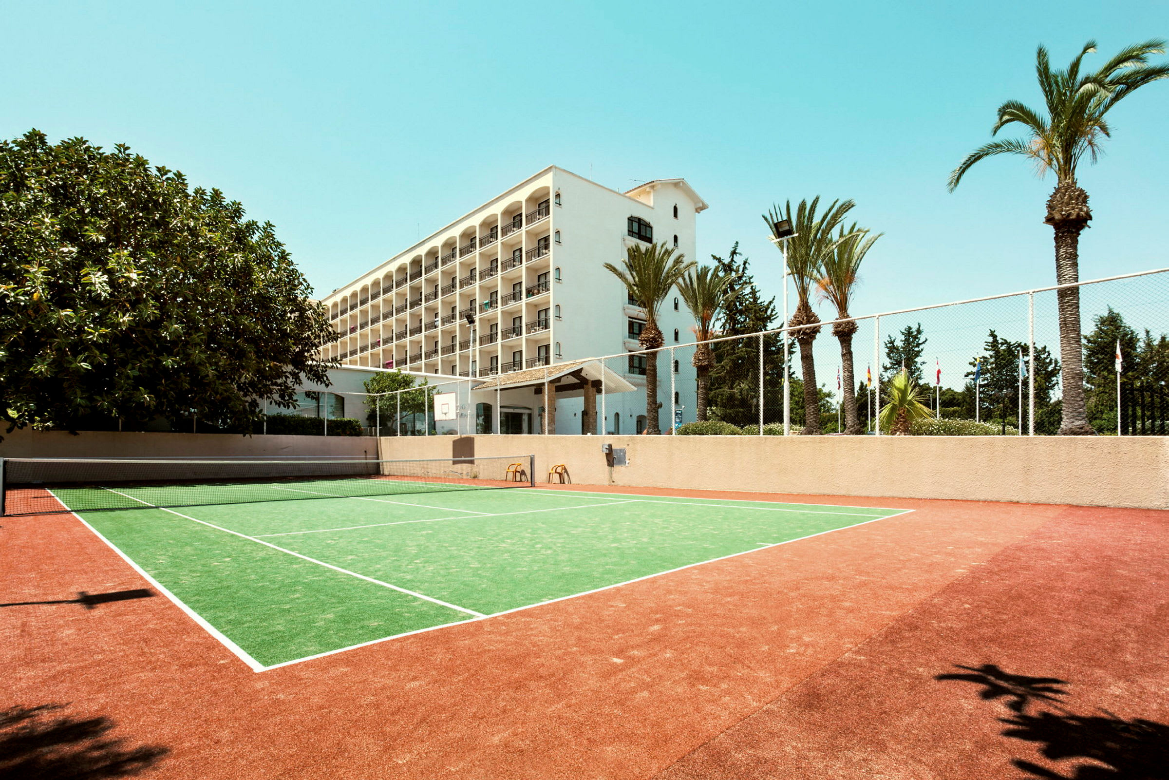 En tennisbane foran et hotel med flere etager og omgivet af palmer. (AI-genereret tekst)