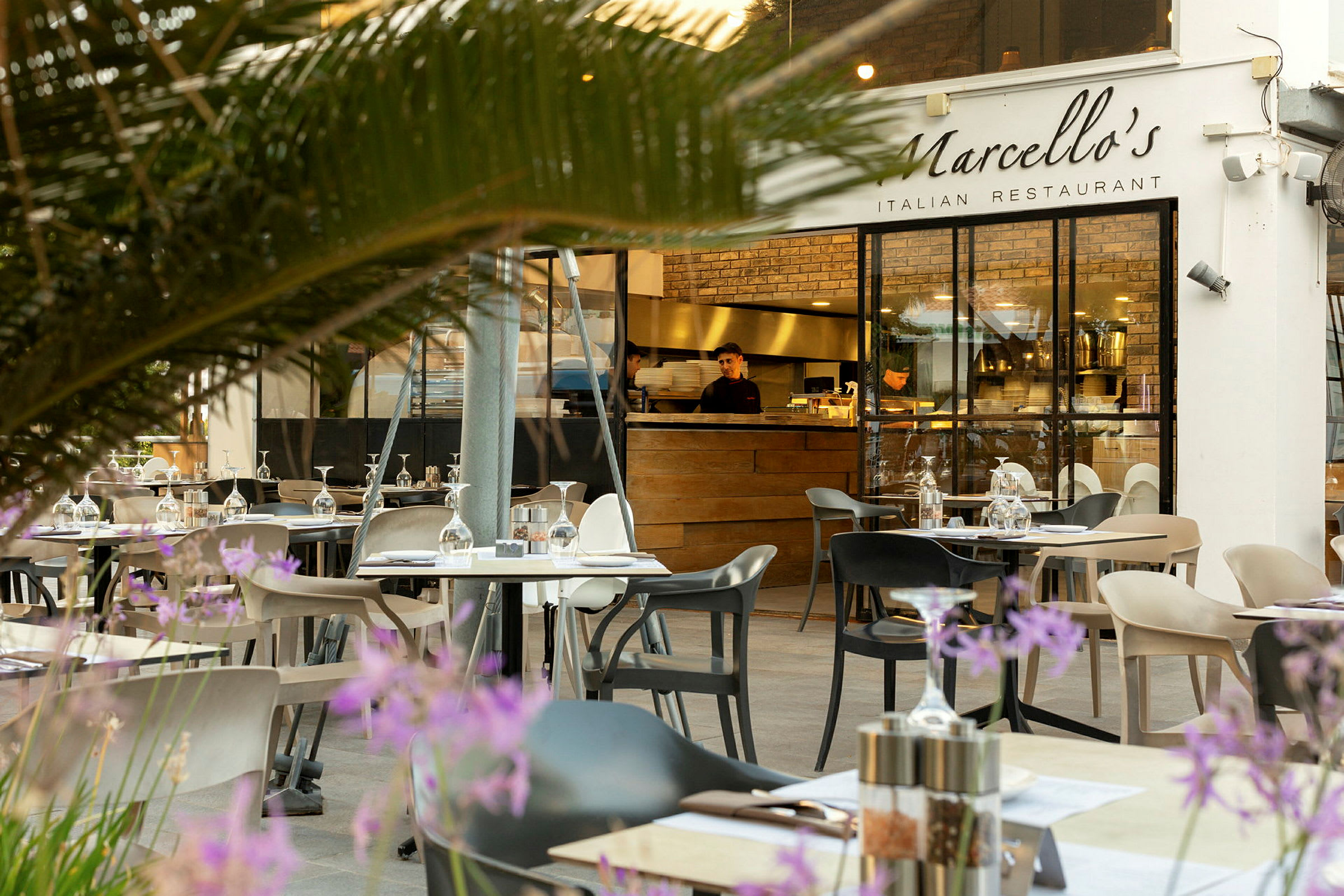 Restaurant med udendørsborde og frodighed i forgrunden. (AI-genereret tekst)