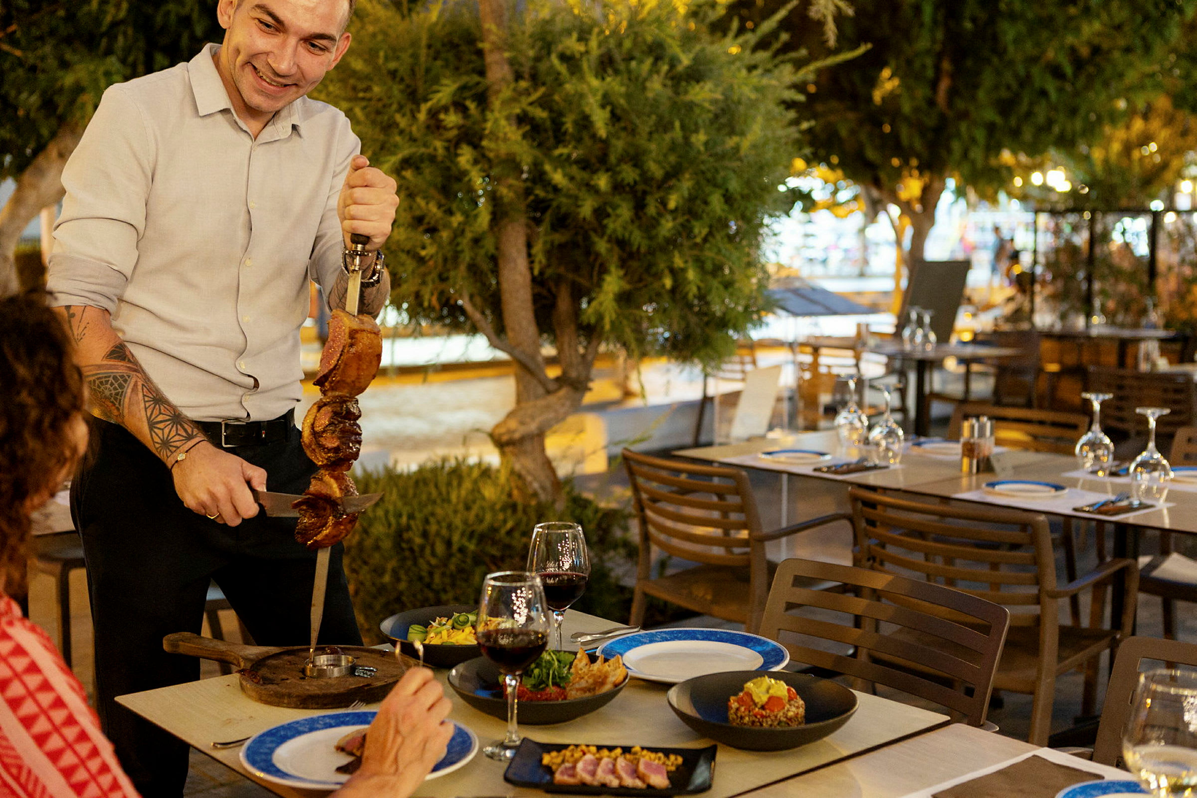 En person serverer grillet kød ved et bord udendørs på en restaurant. (AI-genereret tekst)