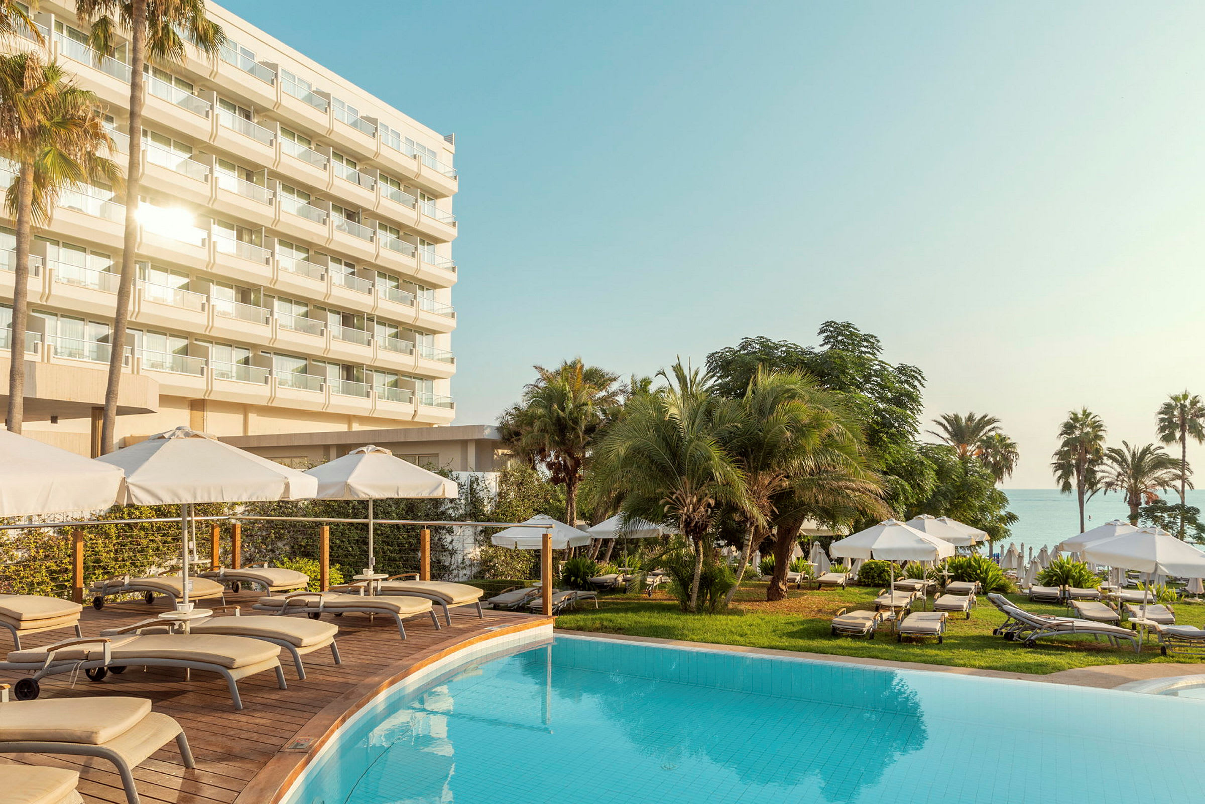 Et hotel med flere etager bag en pool og liggestole omgivet af parasoller og palmer. (AI-genereret tekst)