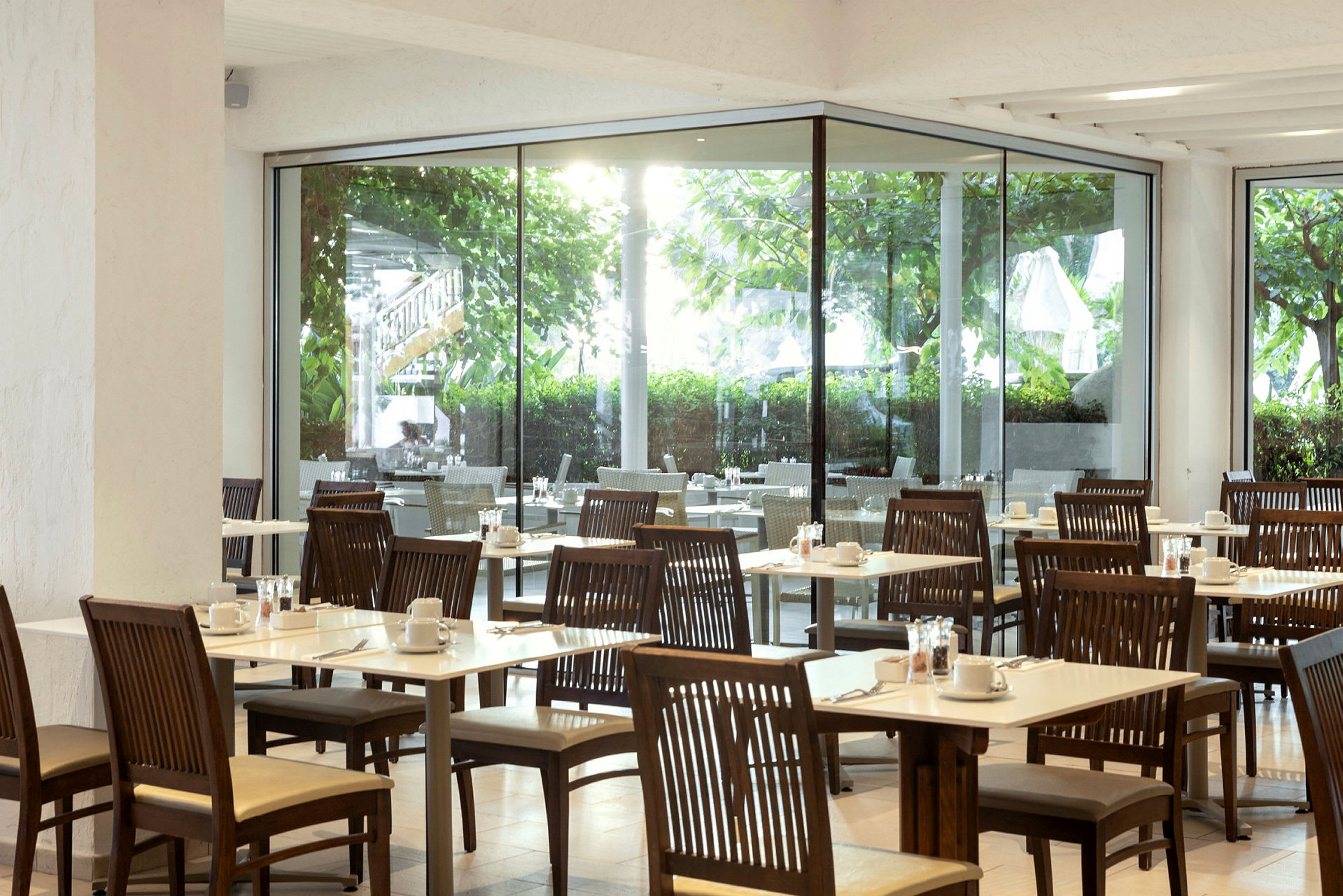 Billede af en restaurant med træborde og stole samt udsigt til grønne omgivelser gennem store glasvinduer. (AI-genereret tekst)