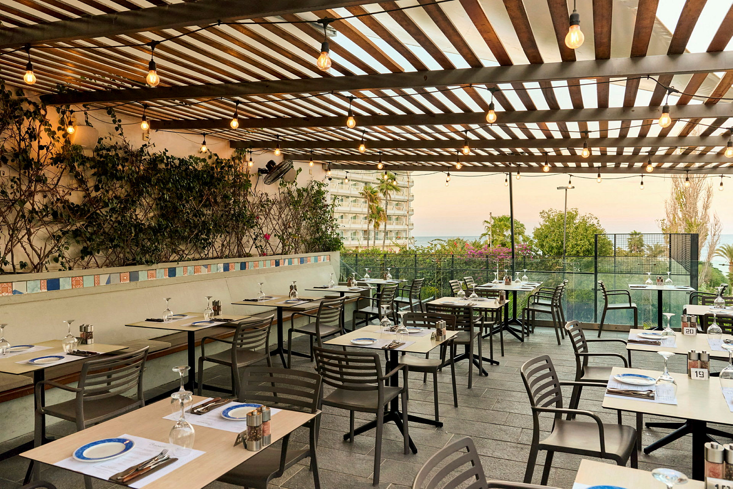 En udendørs restaurant med dækkede borde og stole under en træpergola dekoreret med planter og lyskæder. Udsigt over en bygning og have mod havet. (AI-genereret tekst)