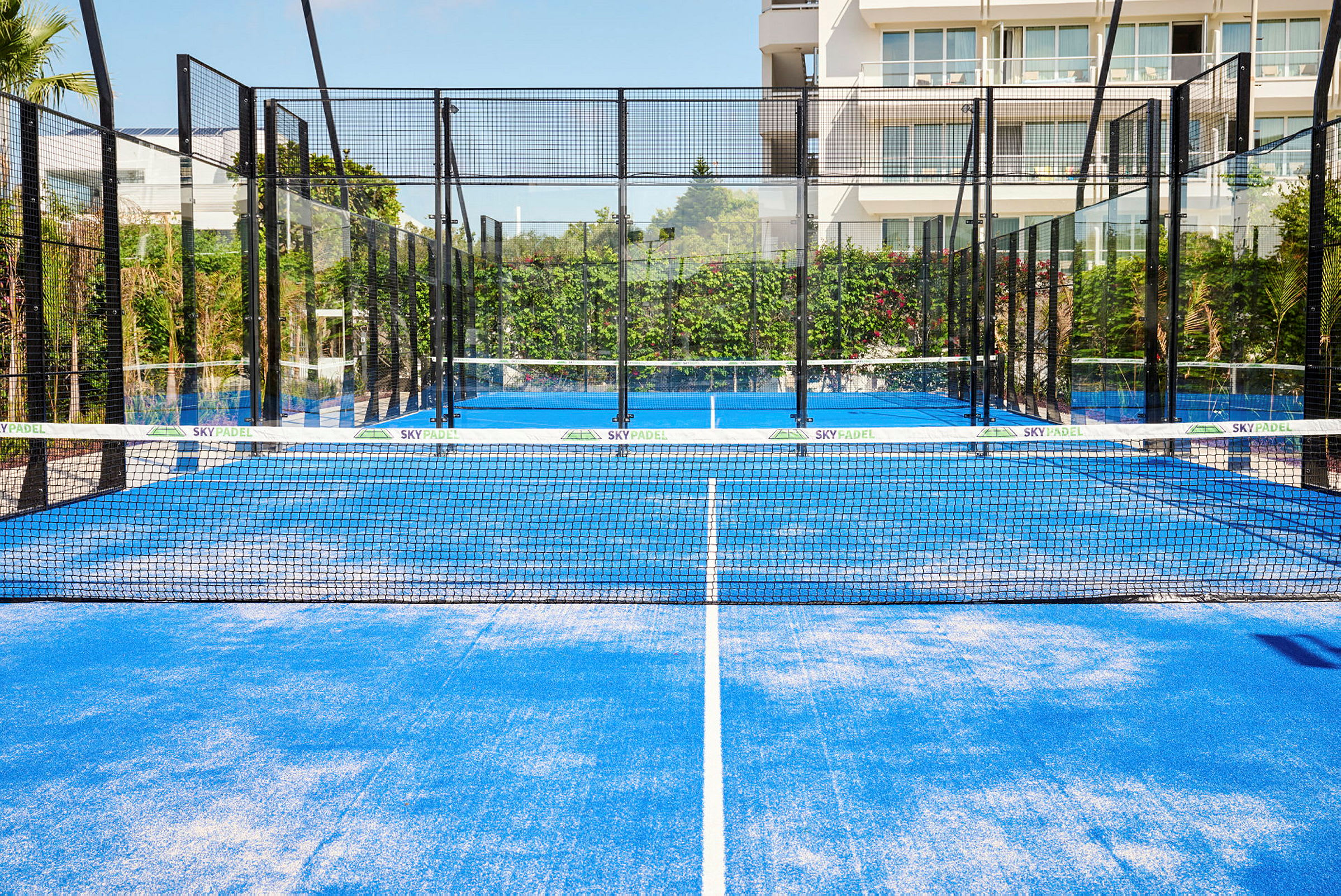 En blå padelbane med net og omgivelser af glasvægge, placeret udendørs med beplantning og en bygning i baggrunden. (AI-genereret tekst)