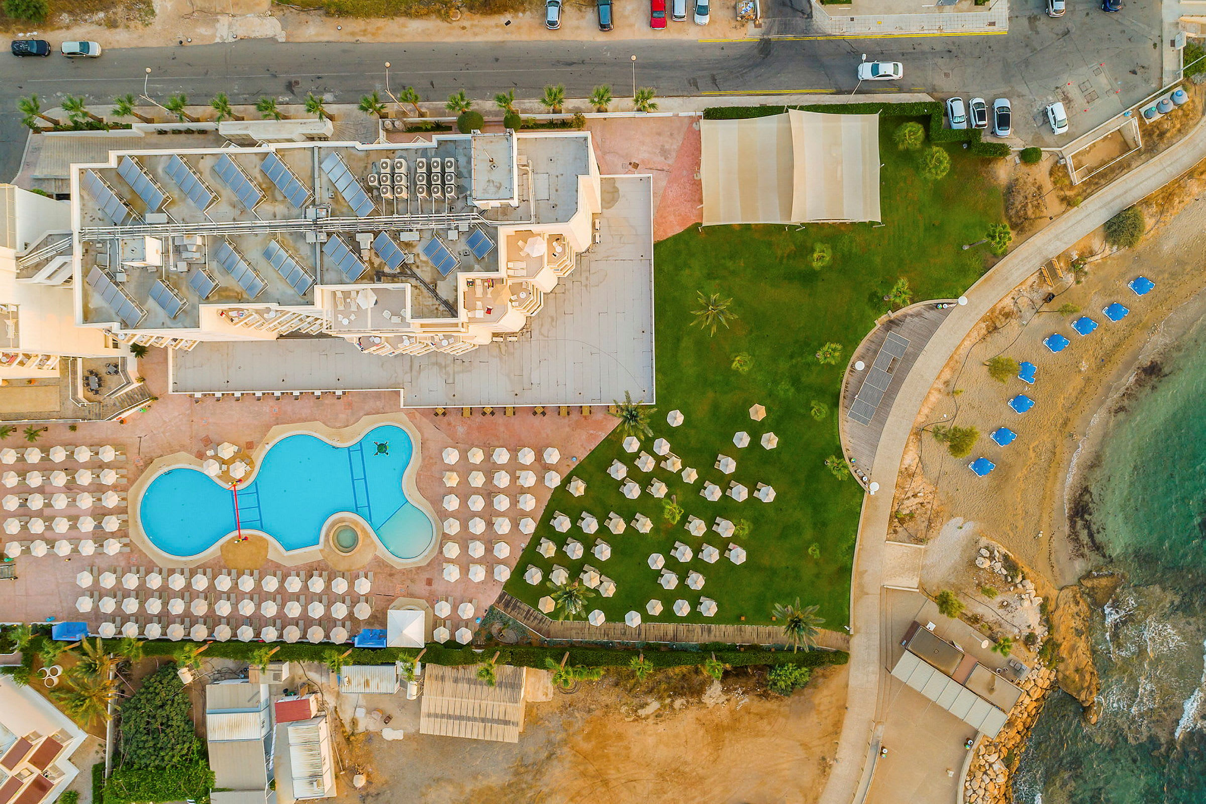 Luftfoto af hotelområdet med swimmingpool, solsenge, græsareal og strand med parasoller. (AI-genereret tekst)
