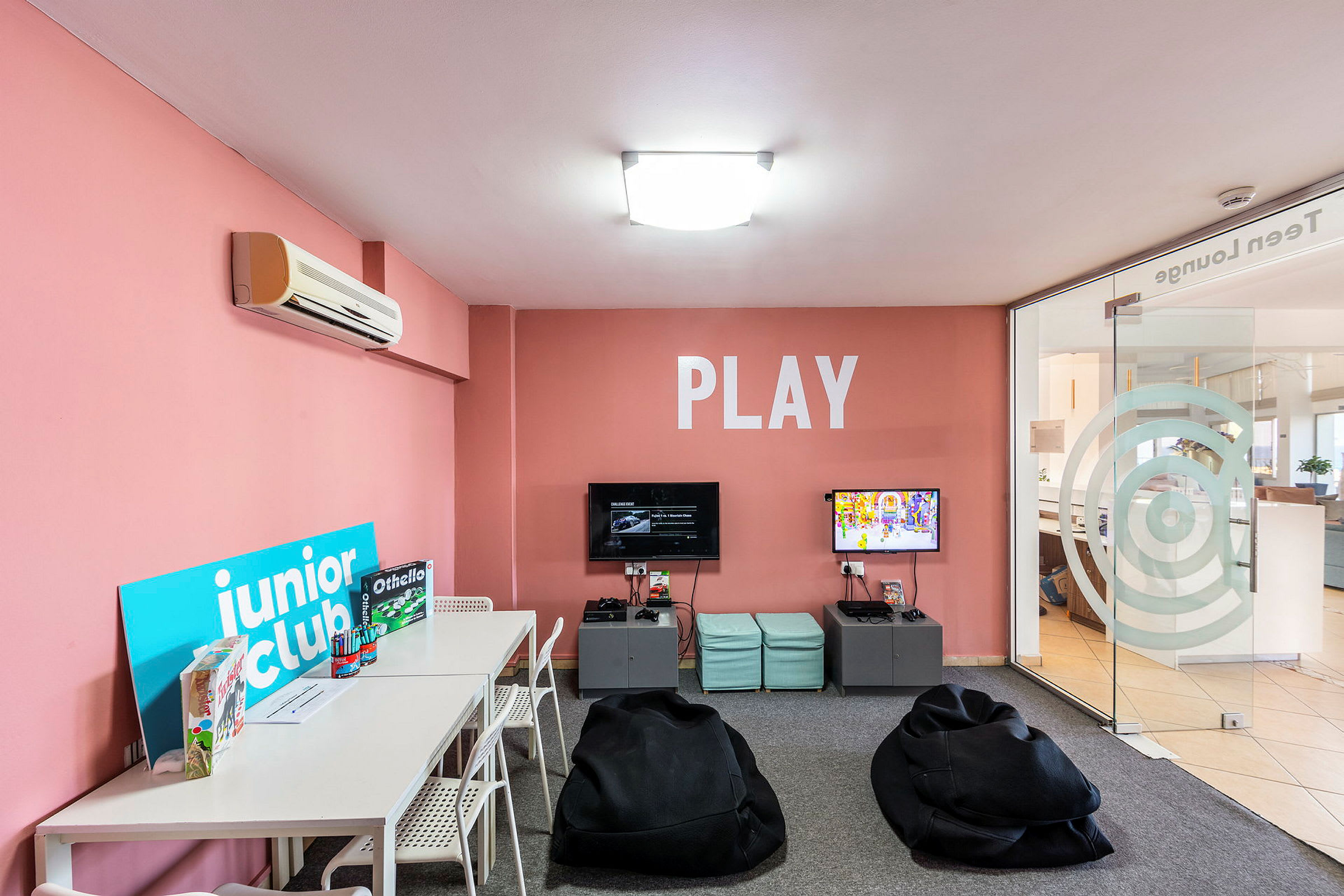 Et legerum med lyserøde vægge, borde, stole og spil samt sækkestole, og teksten 'PLAY' på væggen. (AI-genereret tekst)