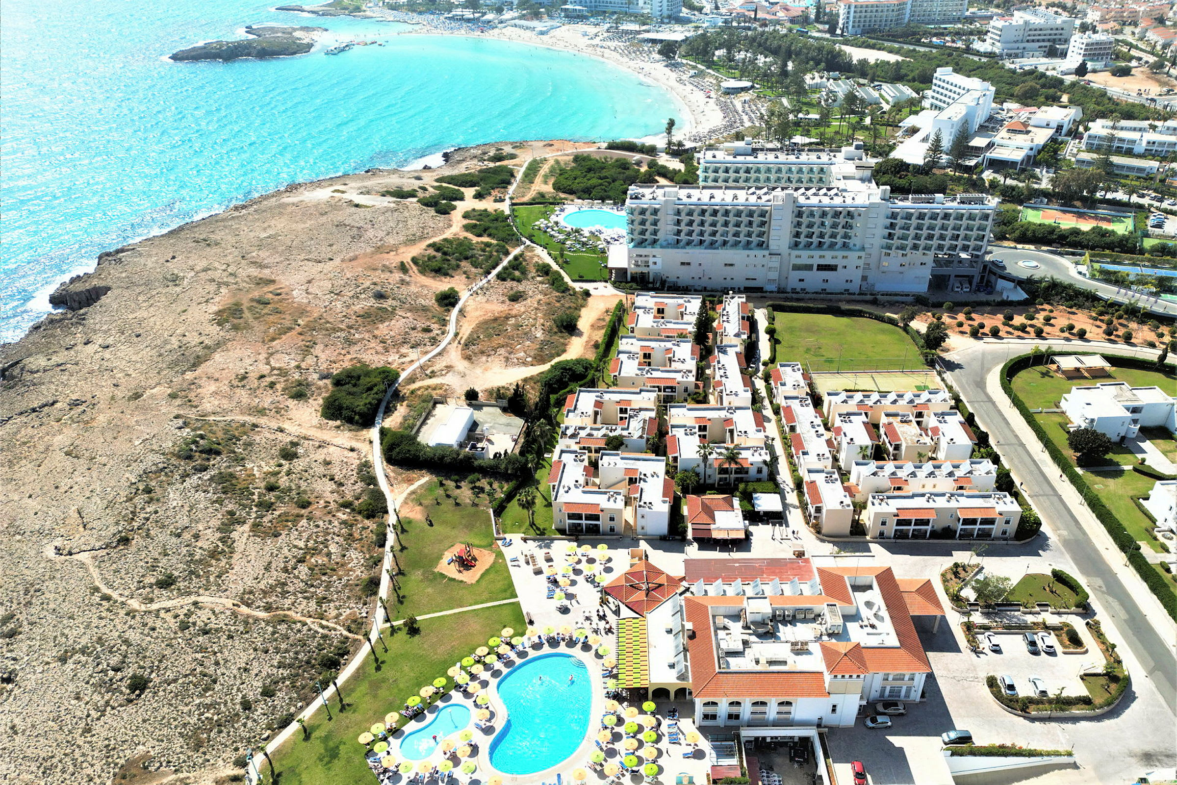 Luftfoto af hotelkomplekset med pool, omkringliggende bygninger og en klippet kyst med udsigt over havet. (AI-genereret tekst)