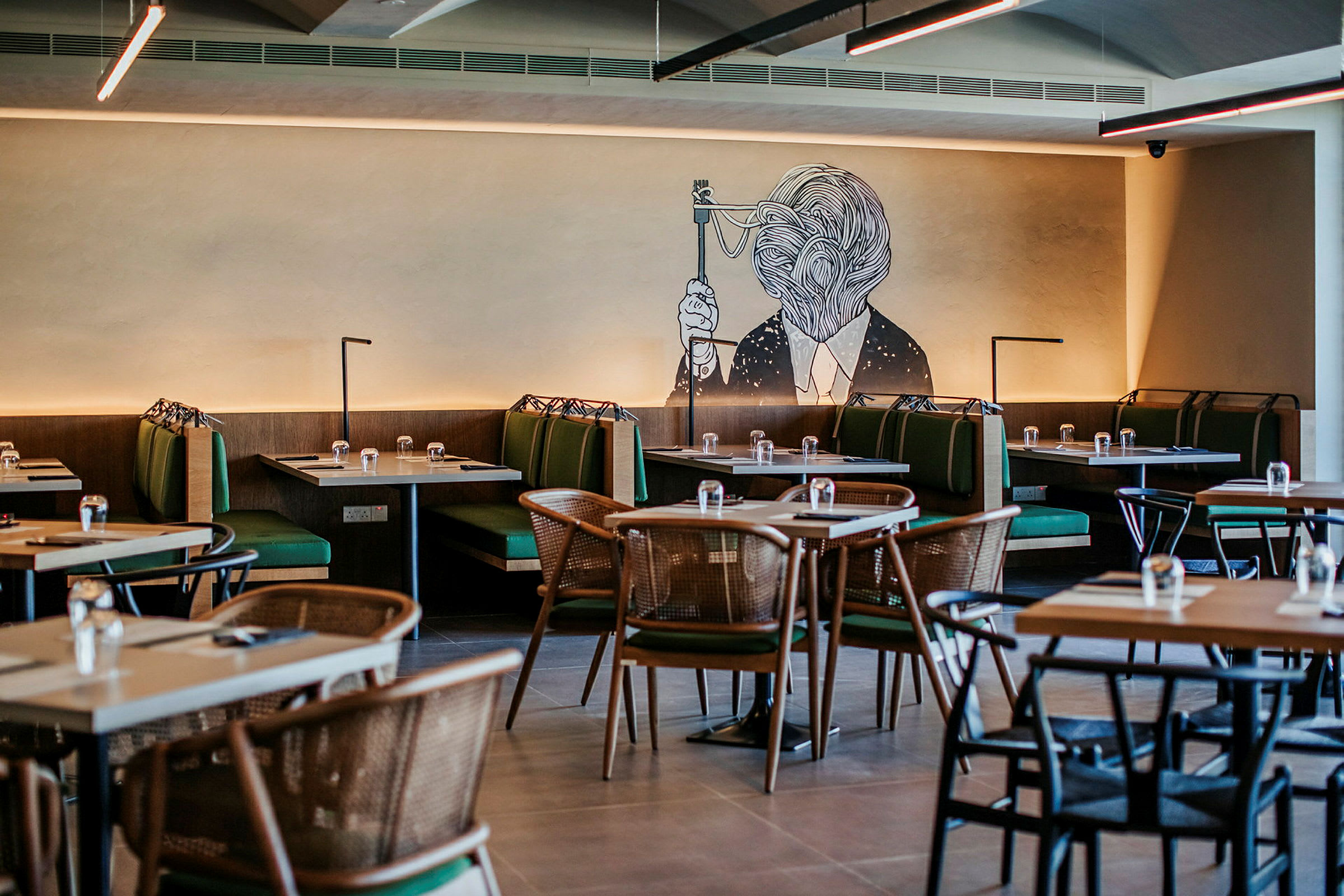 En restaurant med borde og grønne sofaer samt et vægmaleri af et kunstværk. (AI-genereret tekst)