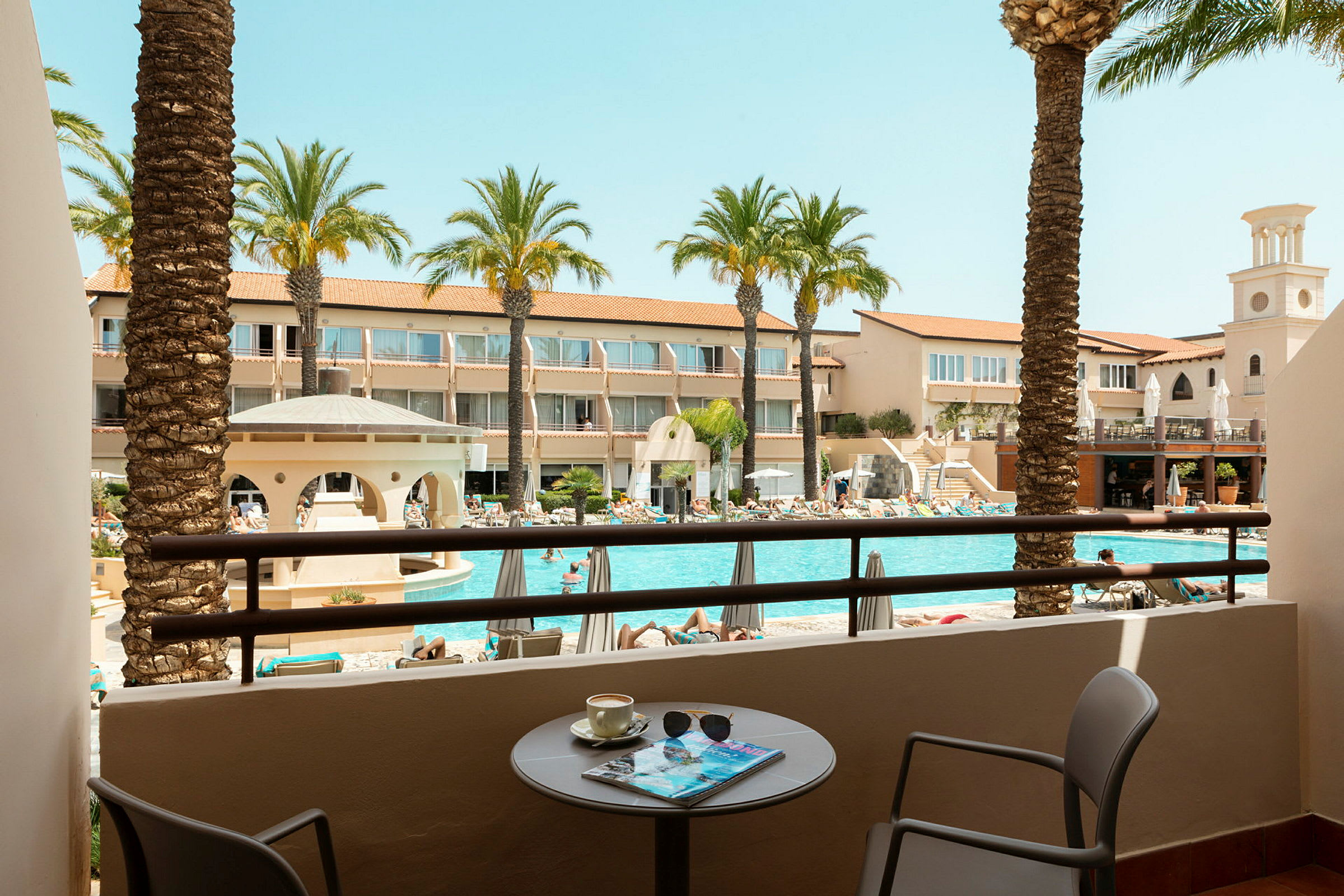Udsigt fra en balkon til en pool og omkringliggende hotelbygninger. (AI-genereret tekst)