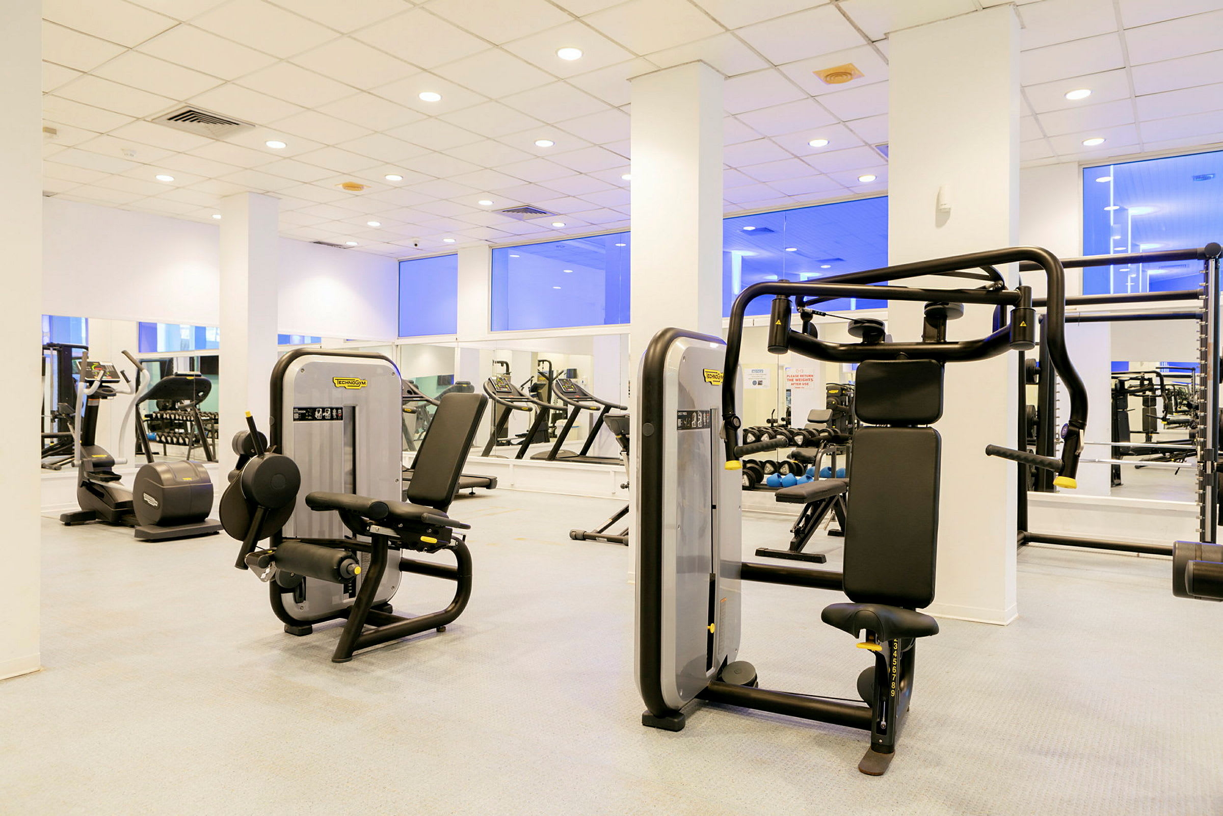 Et fitnesscenter med træningsmaskiner og spejle langs væggene. (AI-genereret tekst)