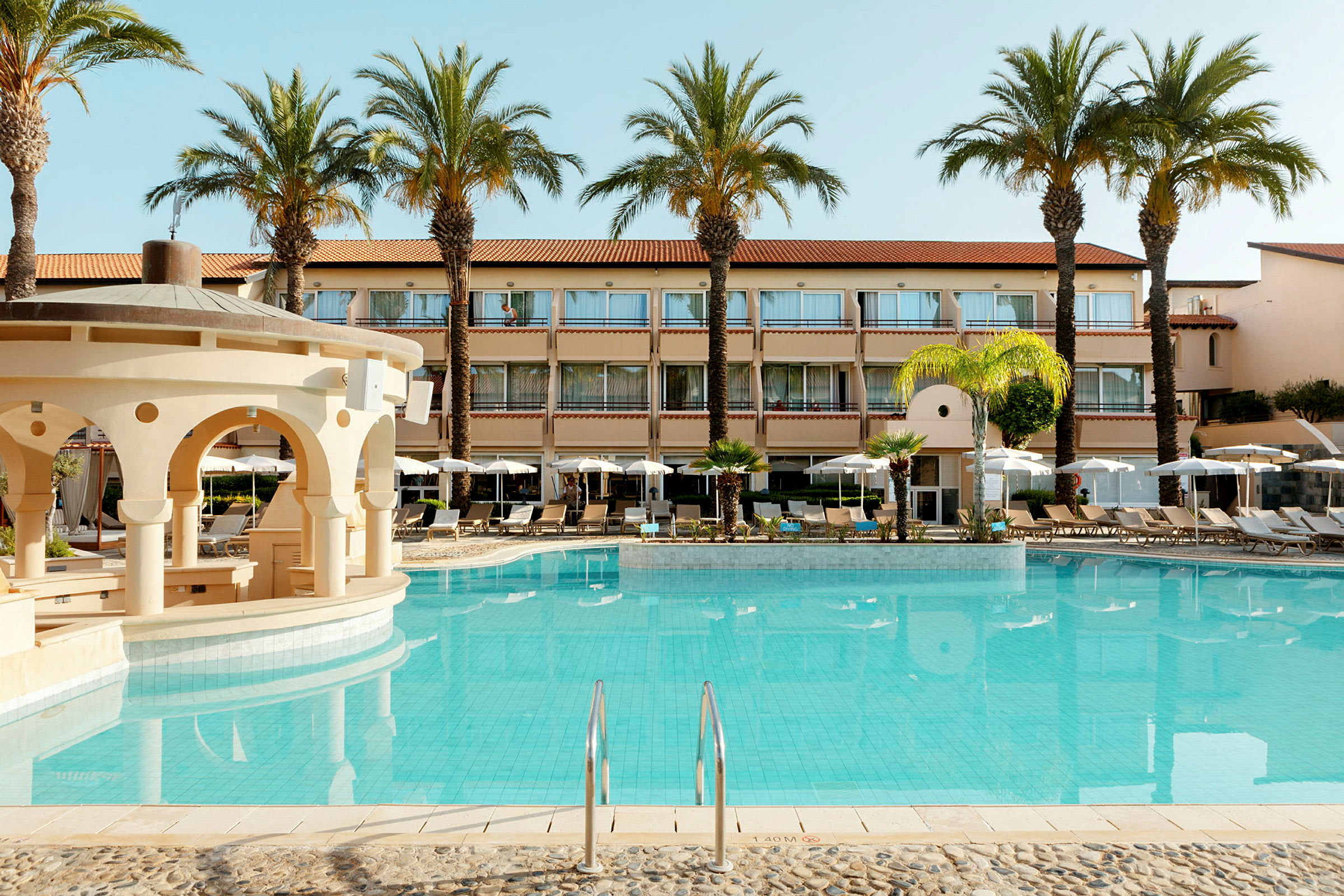 Billede af et hotel med udendørs swimmingpool og omgivende liggestole og parasoller. (AI-genereret tekst)