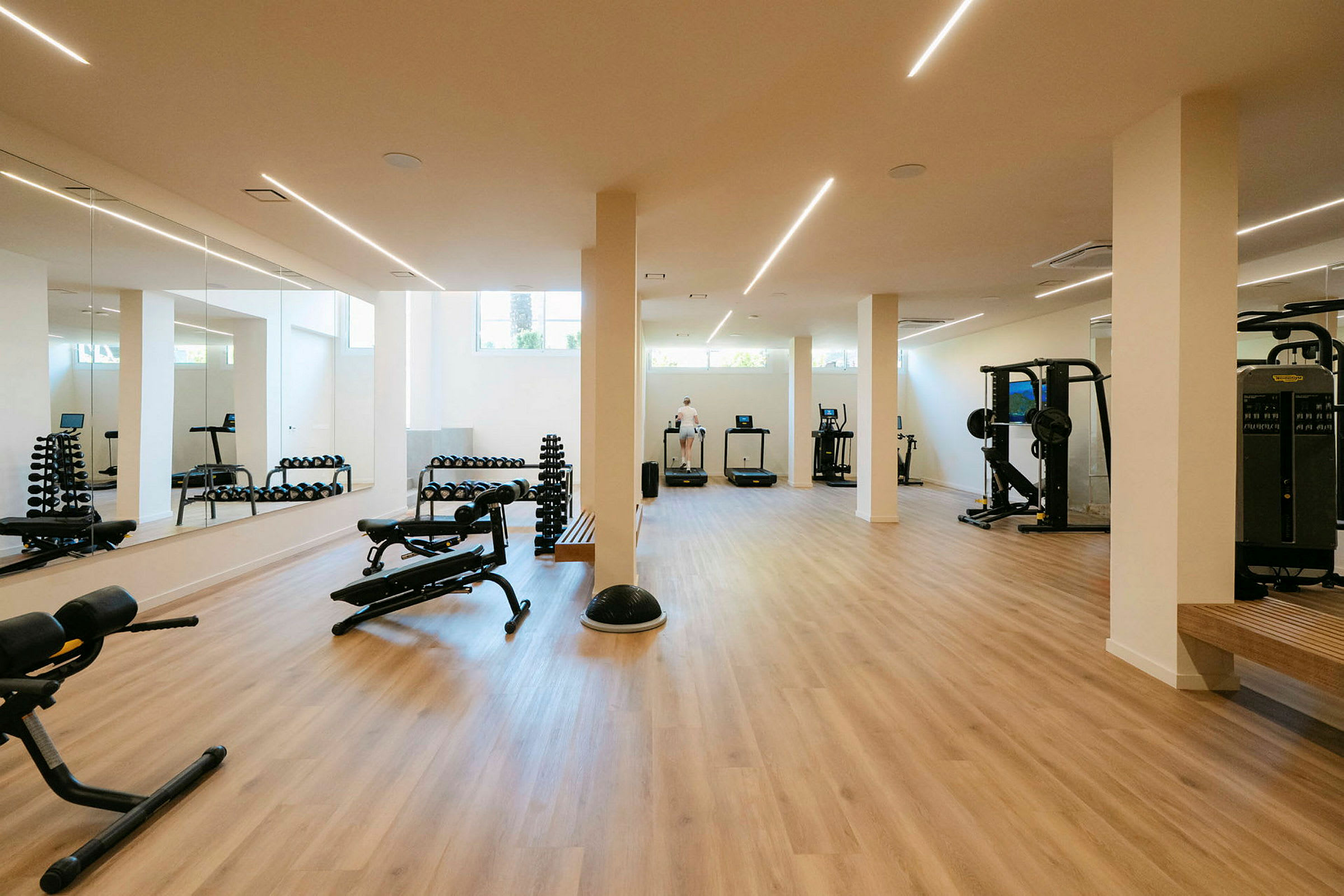Et fitnesscenter med træningsmaskiner og vægte, spejlvægge og lyst trægulv. (AI-genereret tekst)
