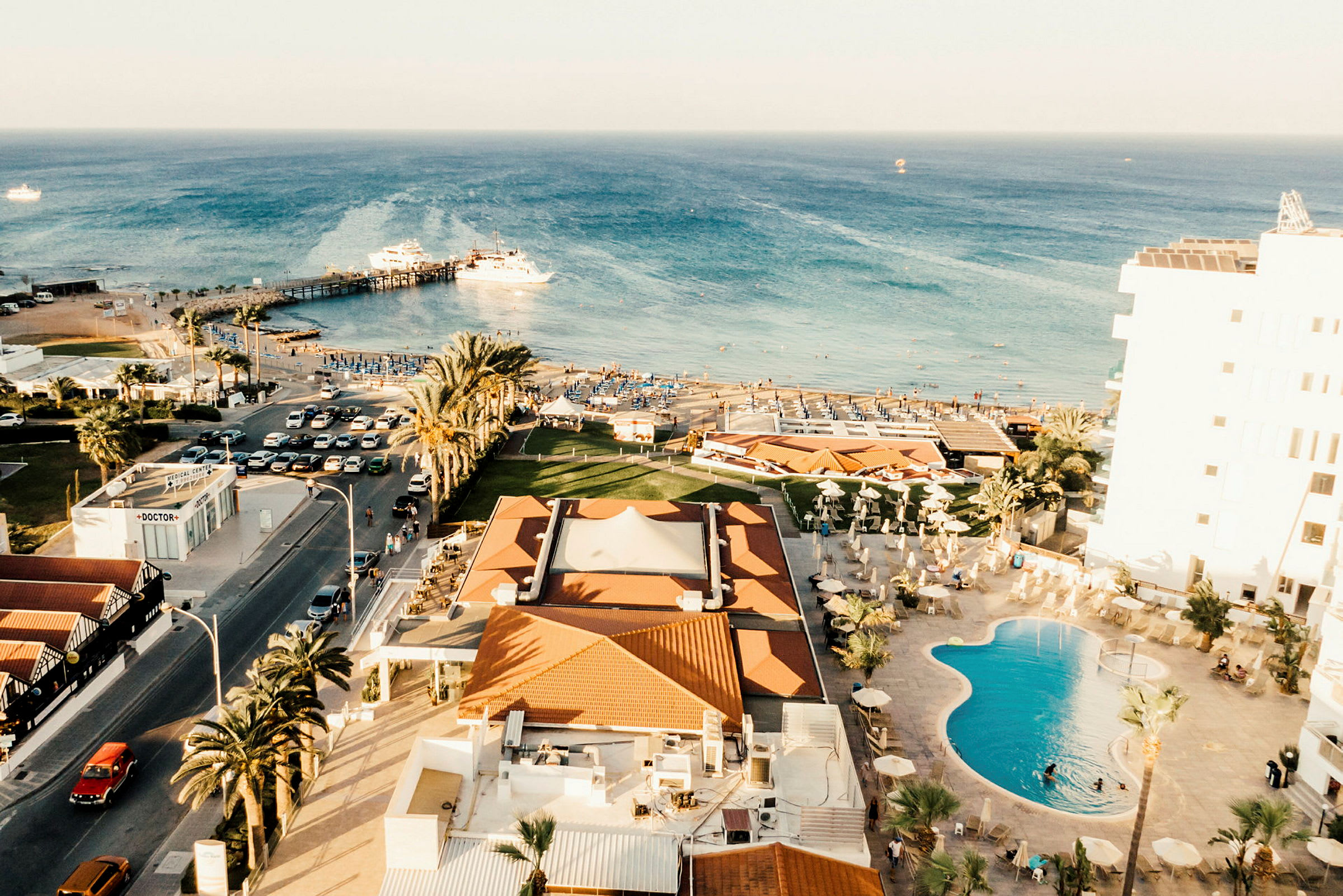 Udsigt over kyst med hotel, pool, palmer og strand, der strækker sig mod havet. (AI-genereret tekst)