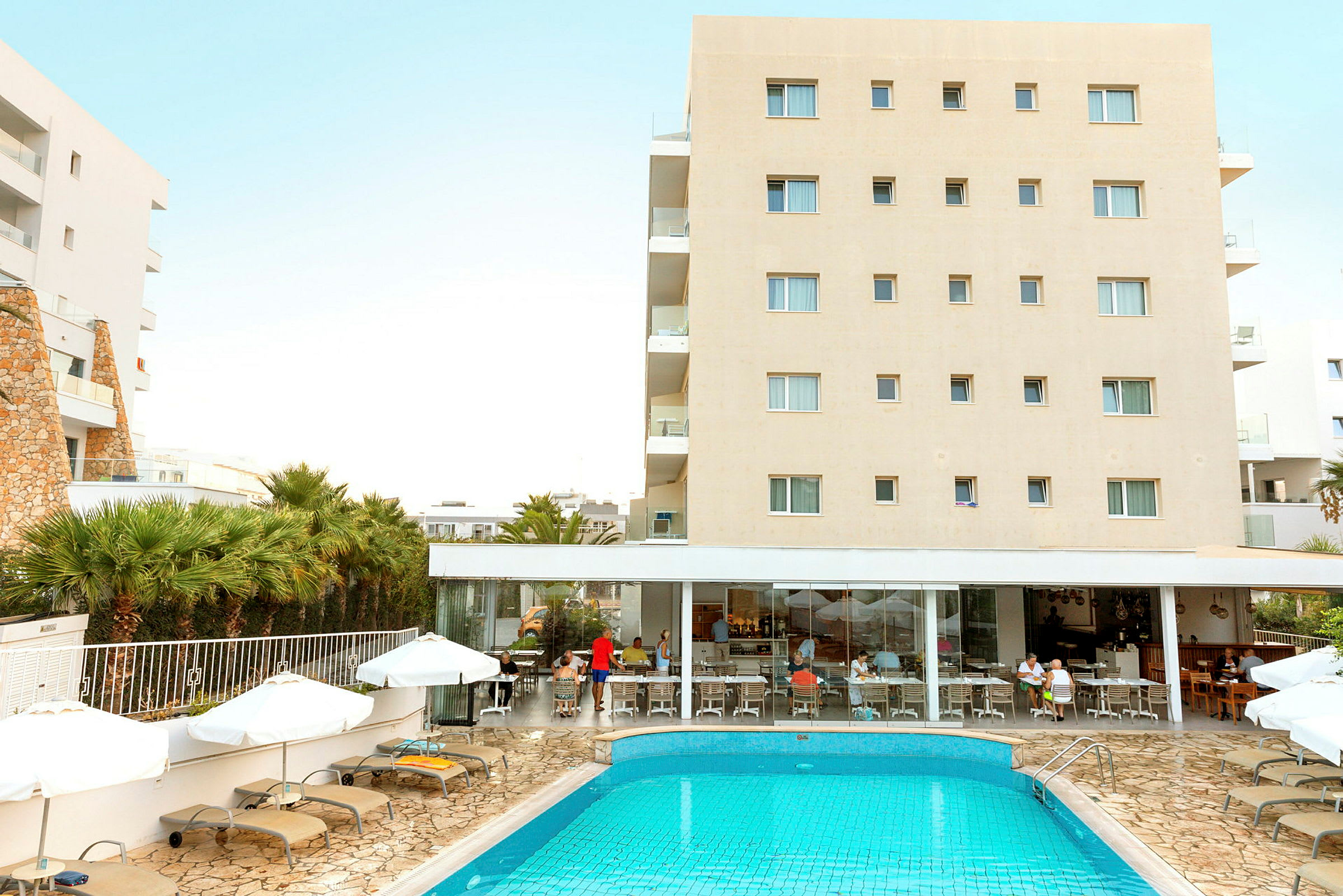 Hotelbygning med swimmingpool og liggestole foran. Folk sidder ved restauranten. (AI-genereret tekst)