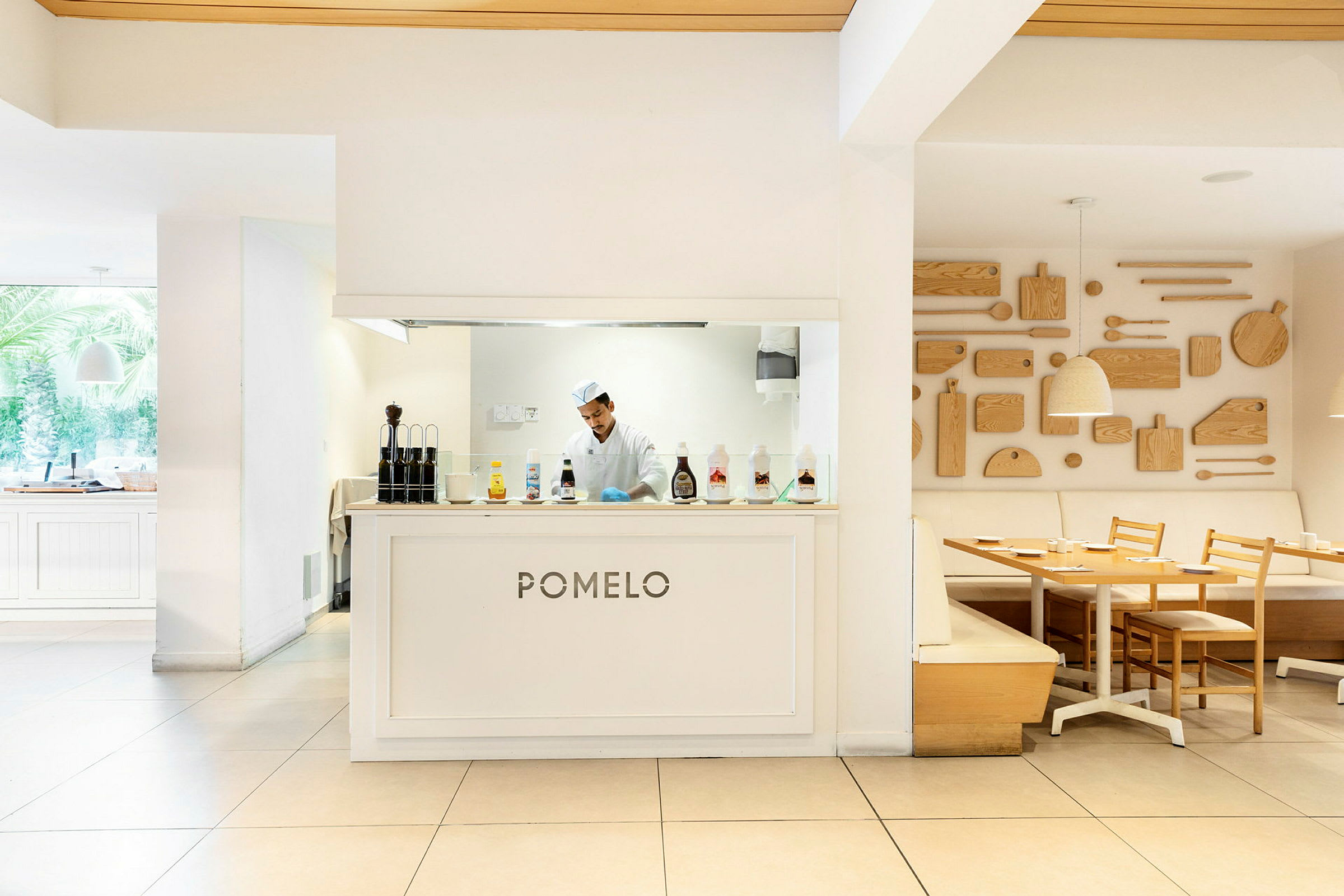 En restaurant med navnet 'Pomelo' og køkkenområde bag disken, møbleret med lyse træborde og stole samt dekorative træpaneler på væggen. (AI-genereret tekst)