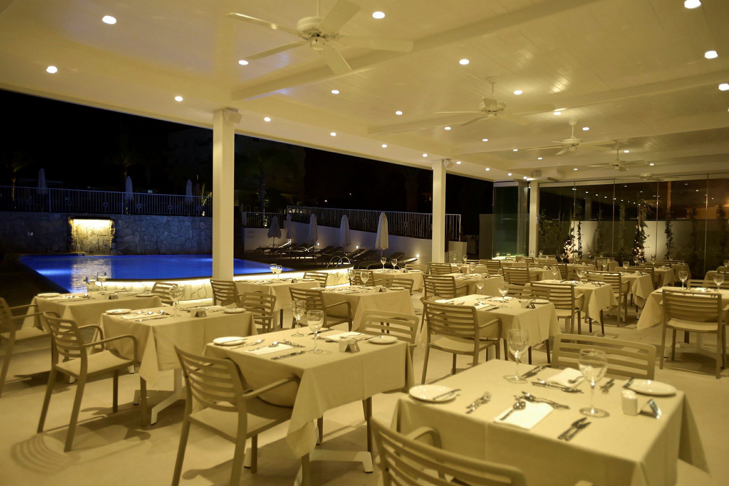 Restaurant med dækkede borde tæt på en swimmingpool om aftenen. (AI-genereret tekst)