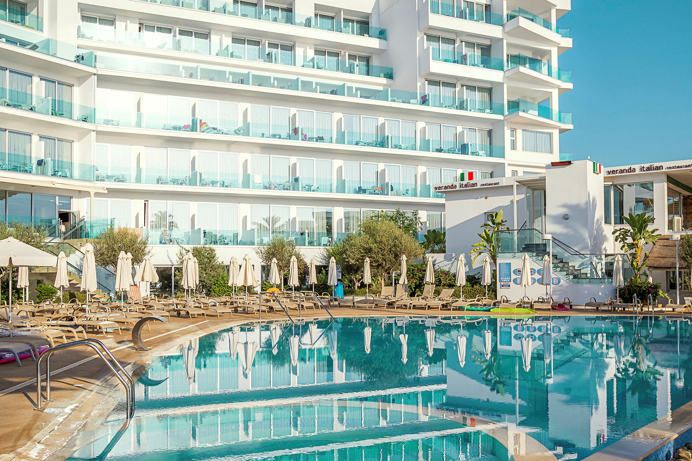 Hotelbygning med balkoner bag en udendørs pool og liggestole. (AI-genereret tekst)