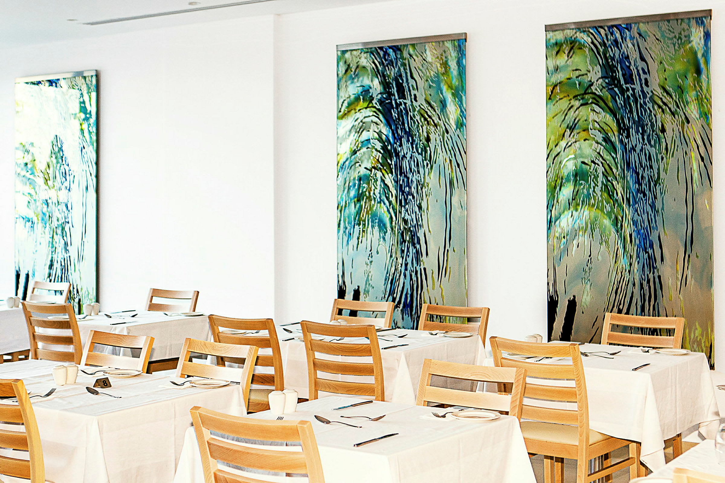 En restaurant med dækkede borde og træstole mod vægge dekoreret med farverige kunstværker. (AI-genereret tekst)