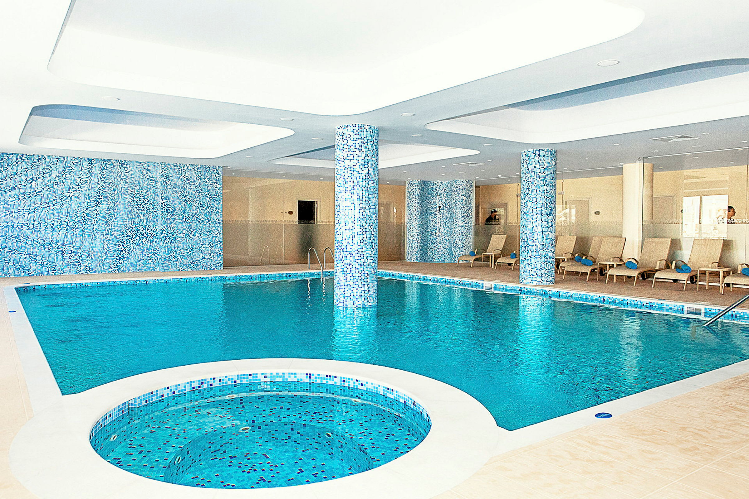 Indendørs swimmingpool med siddeområde og søjler dekoreret med mosaik. (AI-genereret tekst)