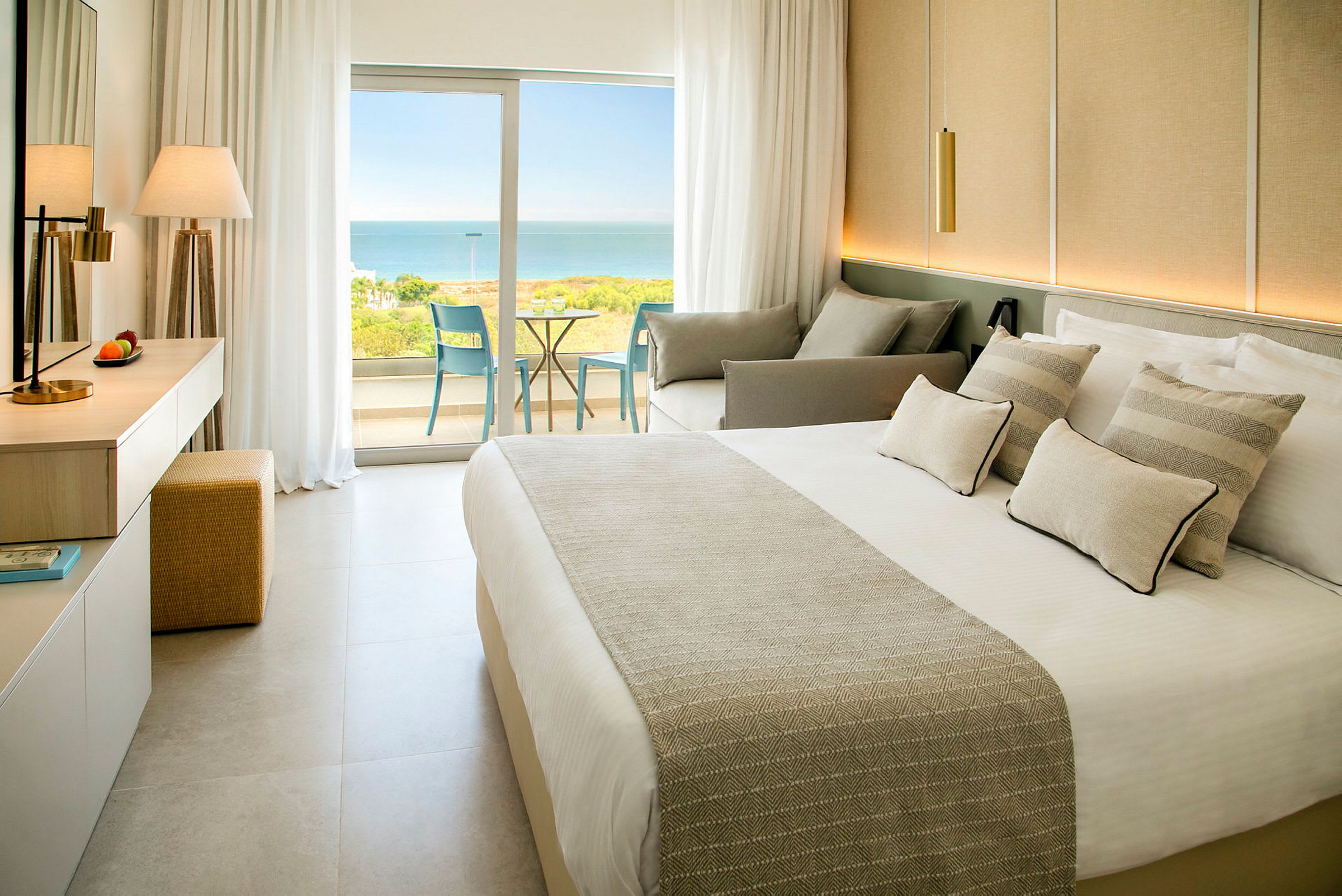 Et hotelværelse med en seng, sofa og glasdøre, der fører ud til en balkon med udsigt over havet. (AI-genereret tekst)