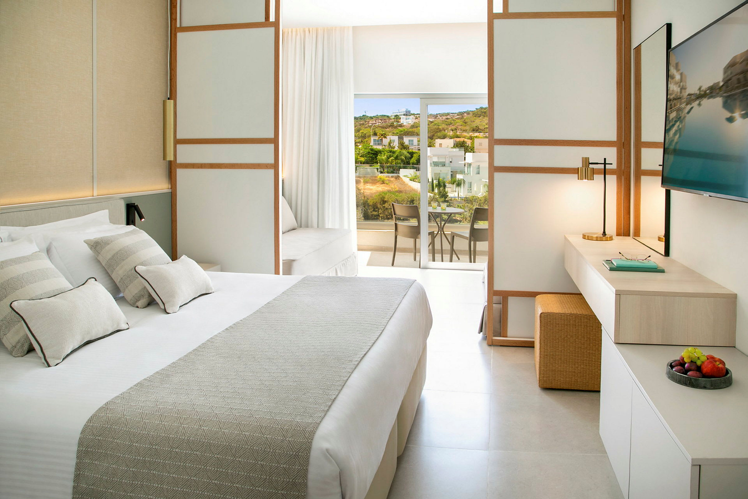 Et lyst hotelværelse med en stor seng, et arbejdsområde og en balkon med udsigt over grønne områder og bygninger. (AI-genereret tekst)