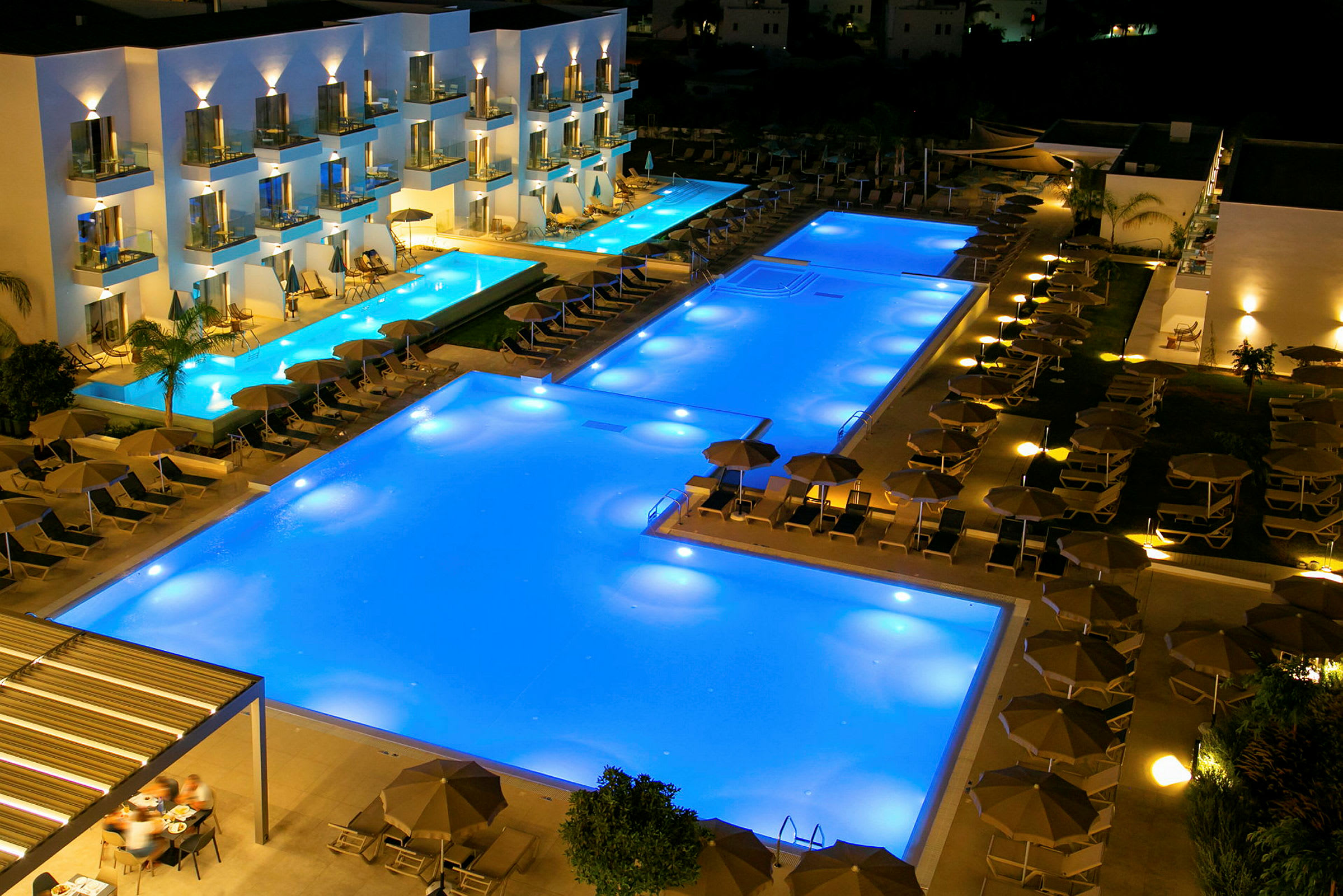Et poolområde ved et hotel om aftenen med oplyste pools og liggestole. (AI-genereret tekst)