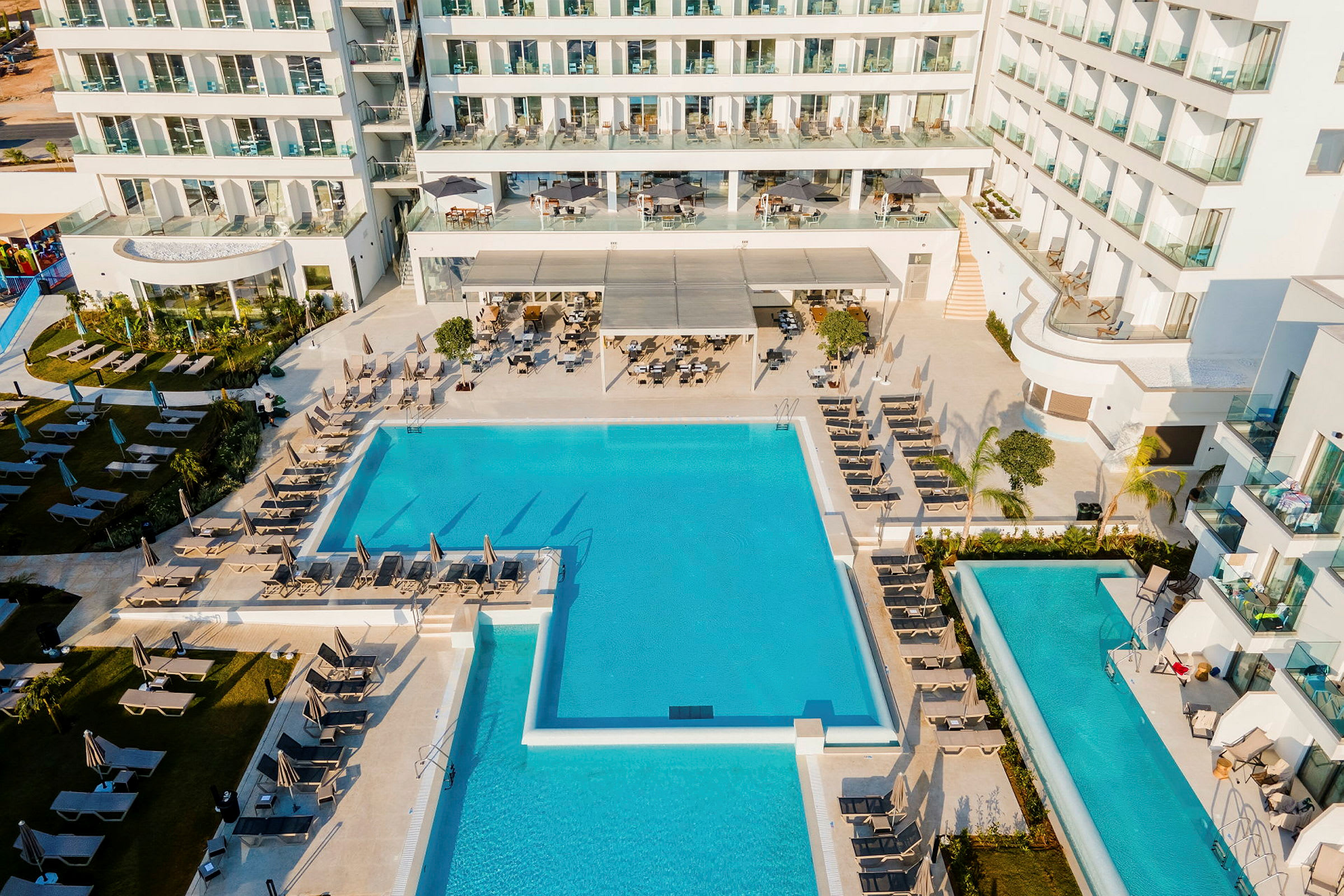 Udsigt over hotellets poolområde med liggestole og facaden af en bygning med balkoner. (AI-genereret tekst)