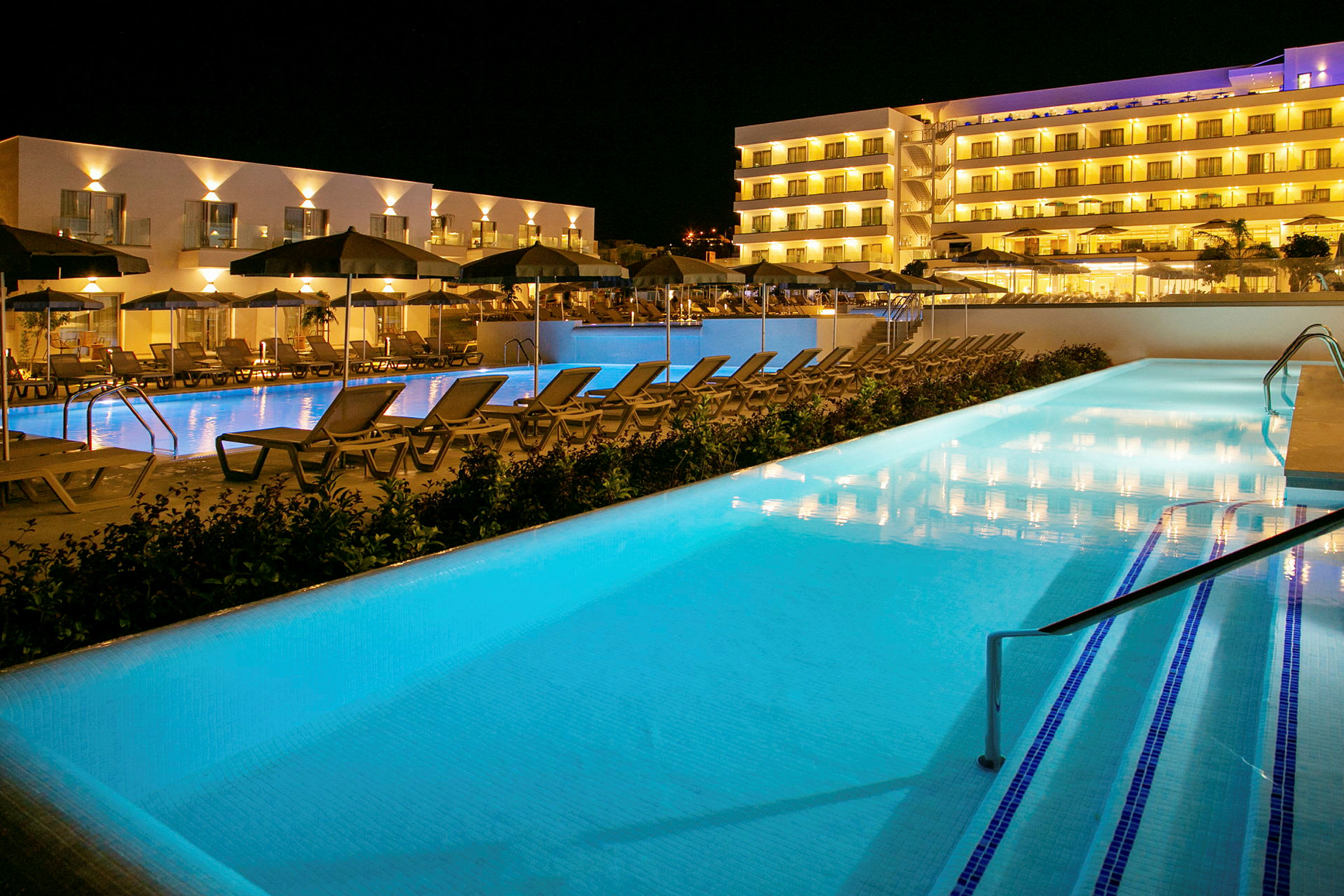 Hotel med oplyste facader og to swimmingpools omgivet af liggestole om aftenen. (AI-genereret tekst)