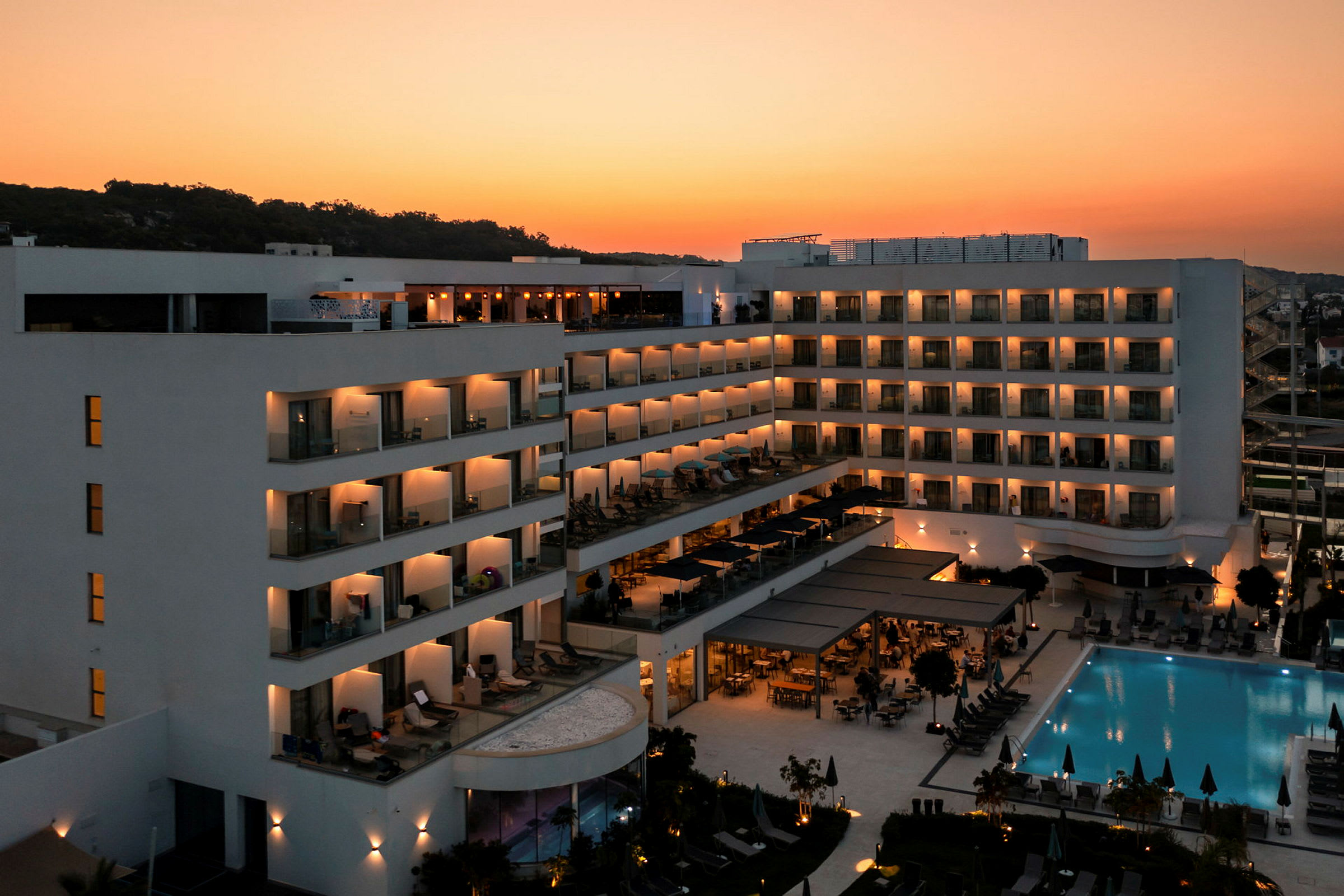 Moderne hotel med oplyste altaner og en swimmingpool ved solnedgang. (AI-genereret tekst)