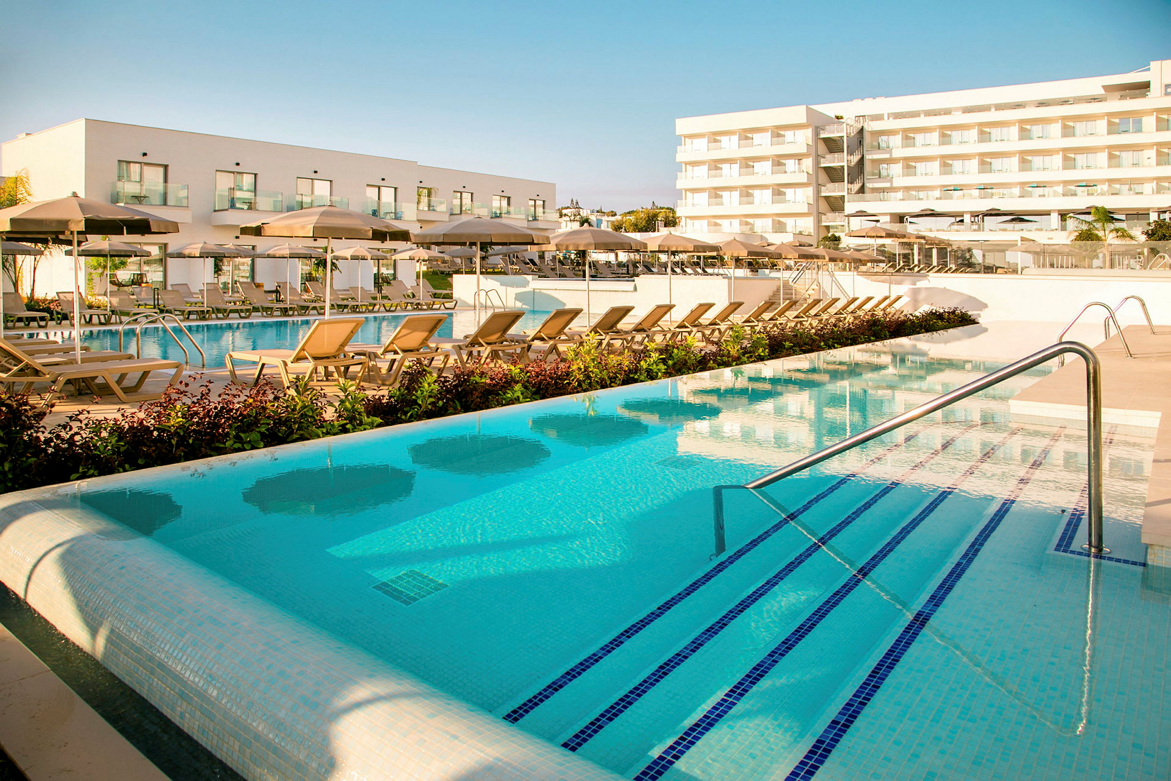 Poolområde med liggestole og parasoller foran moderne hotelbygninger. (AI-genereret tekst)