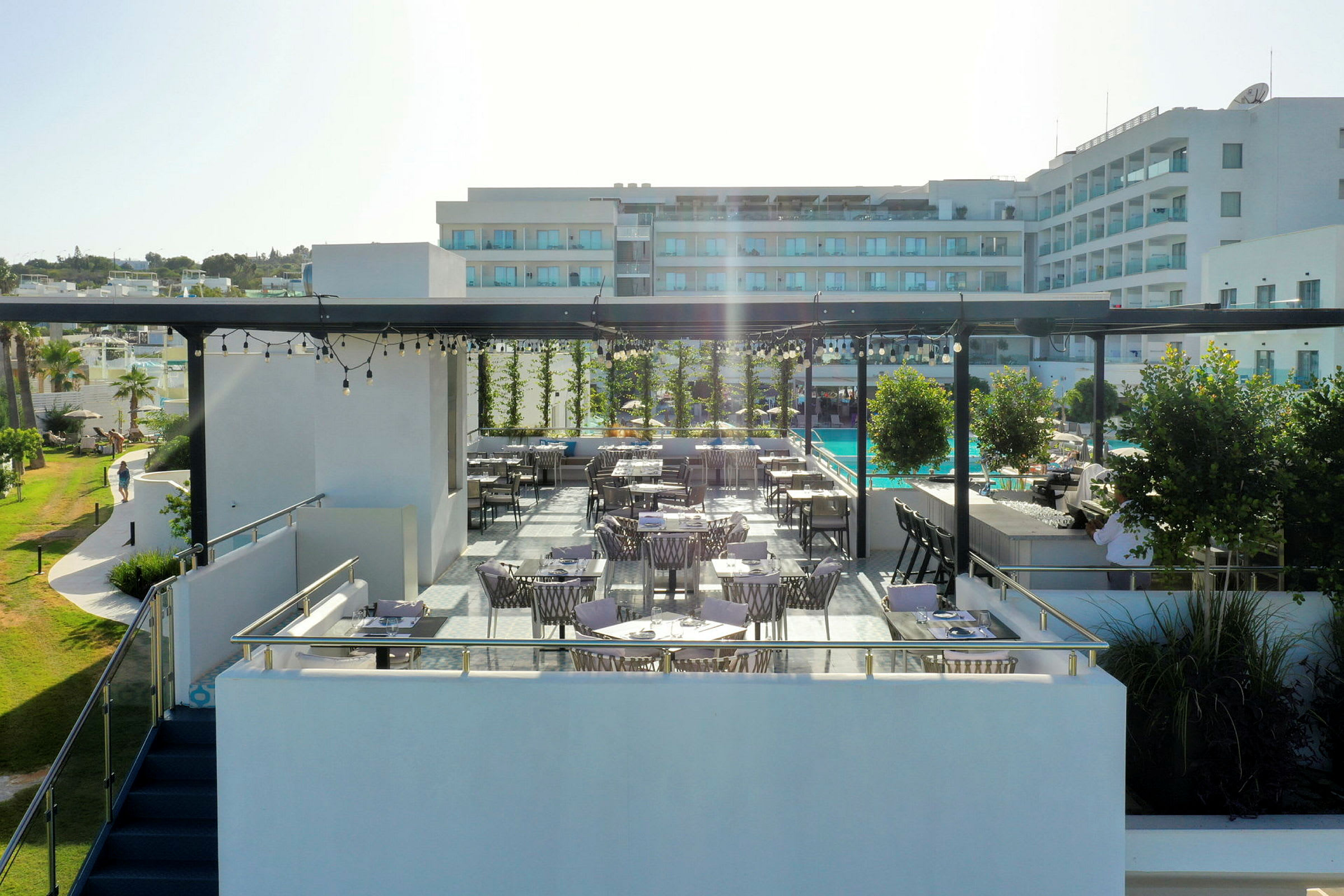 En udendørs terrasse med borde og stole, omgivet af grønne planter og med en hotelbygning i baggrunden. (AI-genereret tekst)