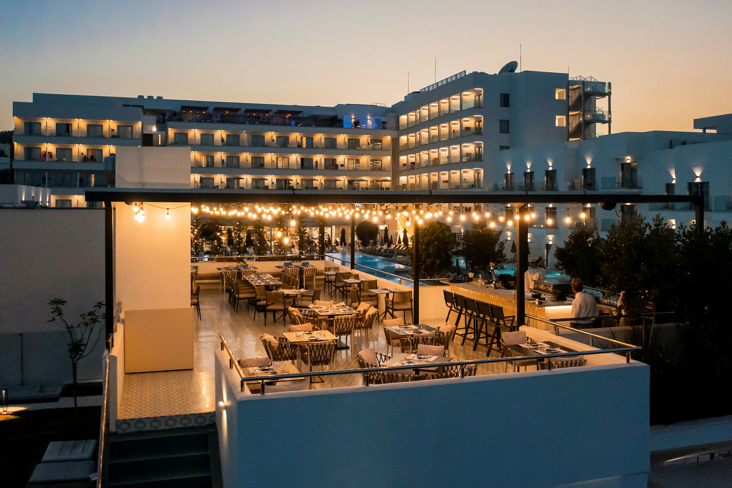 En restaurantterrasse med borde og stole om aftenen, oplyst af lyskæder, med en moderne bygning i baggrunden. (AI-genereret tekst)