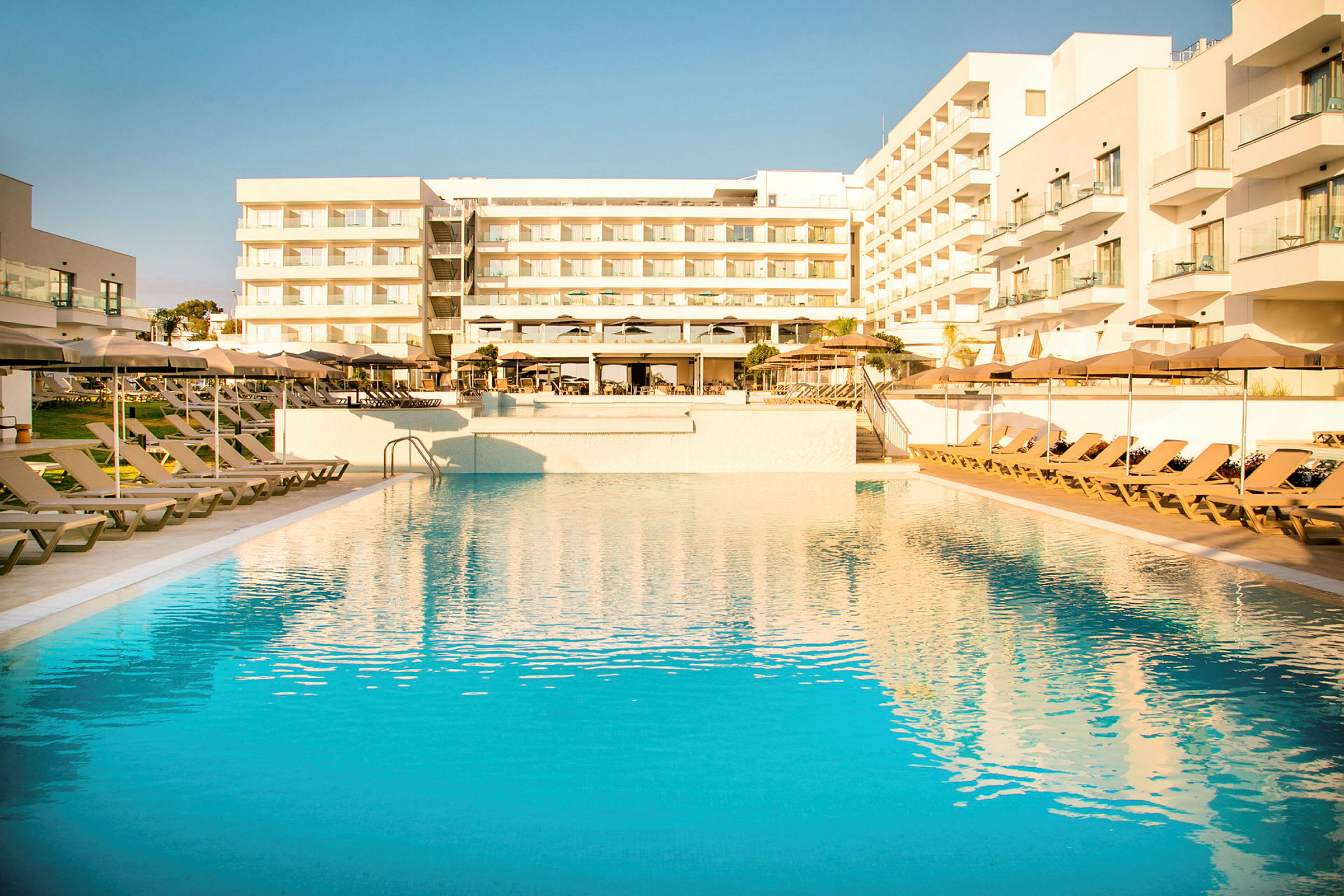 En stor udendørs swimmingpool omgivet af liggestole og parasoller foran et moderne hotel med altaner. (AI-genereret tekst)