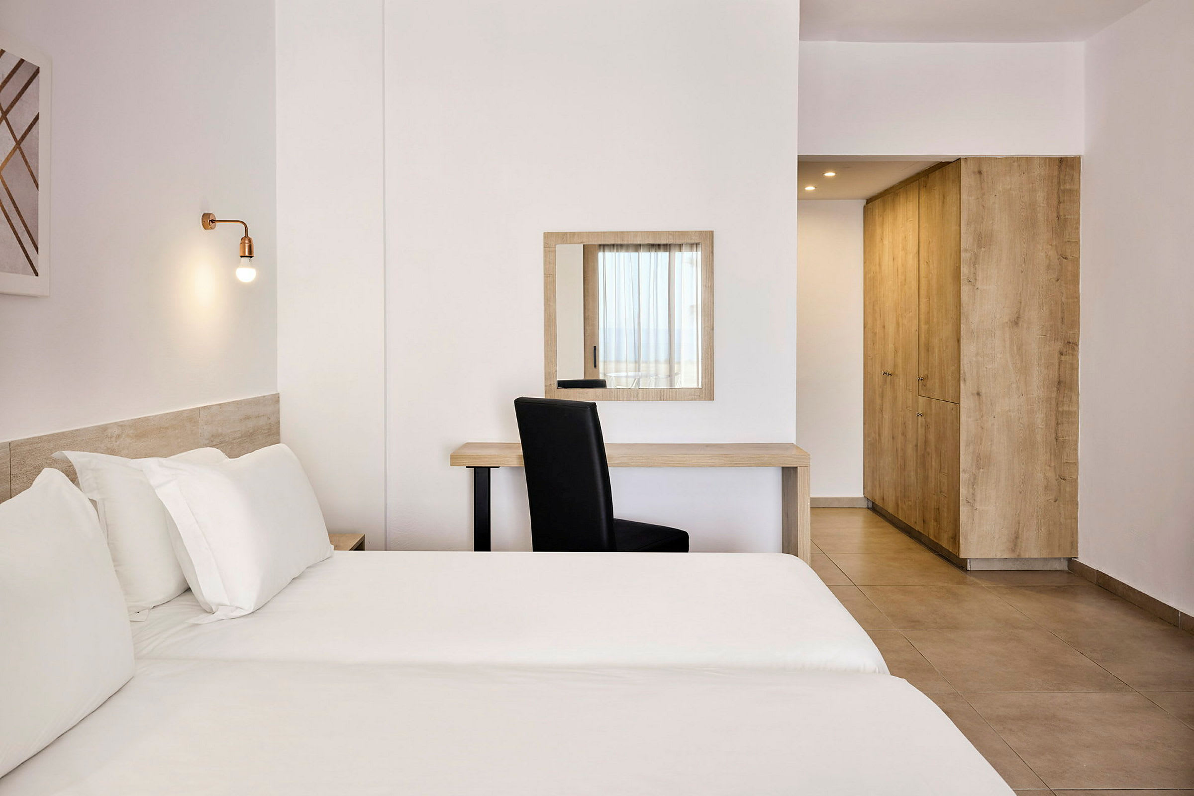 Et lyst hotelværelse med en seng, skrivebord, stol og garderobe. (AI-genereret tekst)