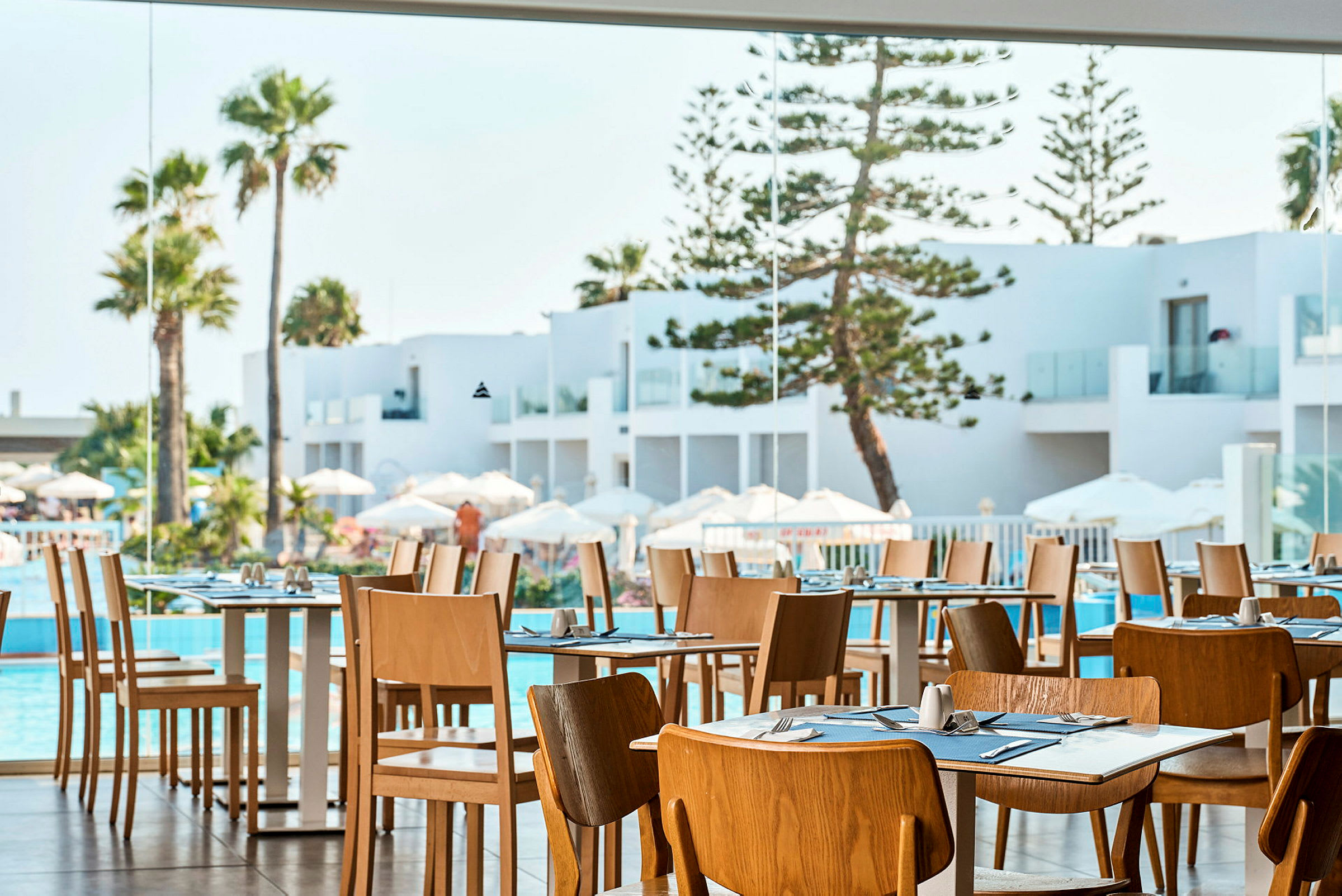 Restaurant med træstole og dækkede borde, udsigt til hvid arkitektur, pool og palmer. (AI-genereret tekst)