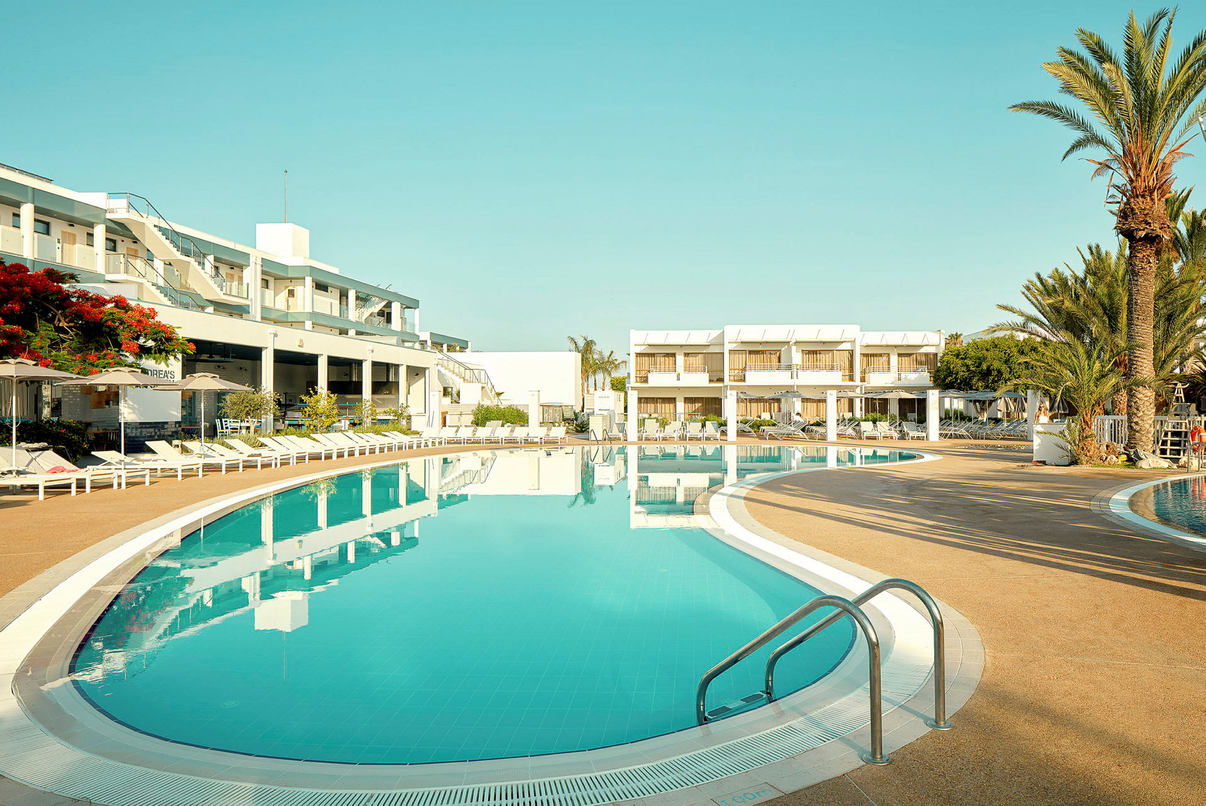 Et hotelområde med en stor pool og omgivede liggestole. I baggrunden er der hotelbygninger og palmetræer. (AI-genereret tekst)
