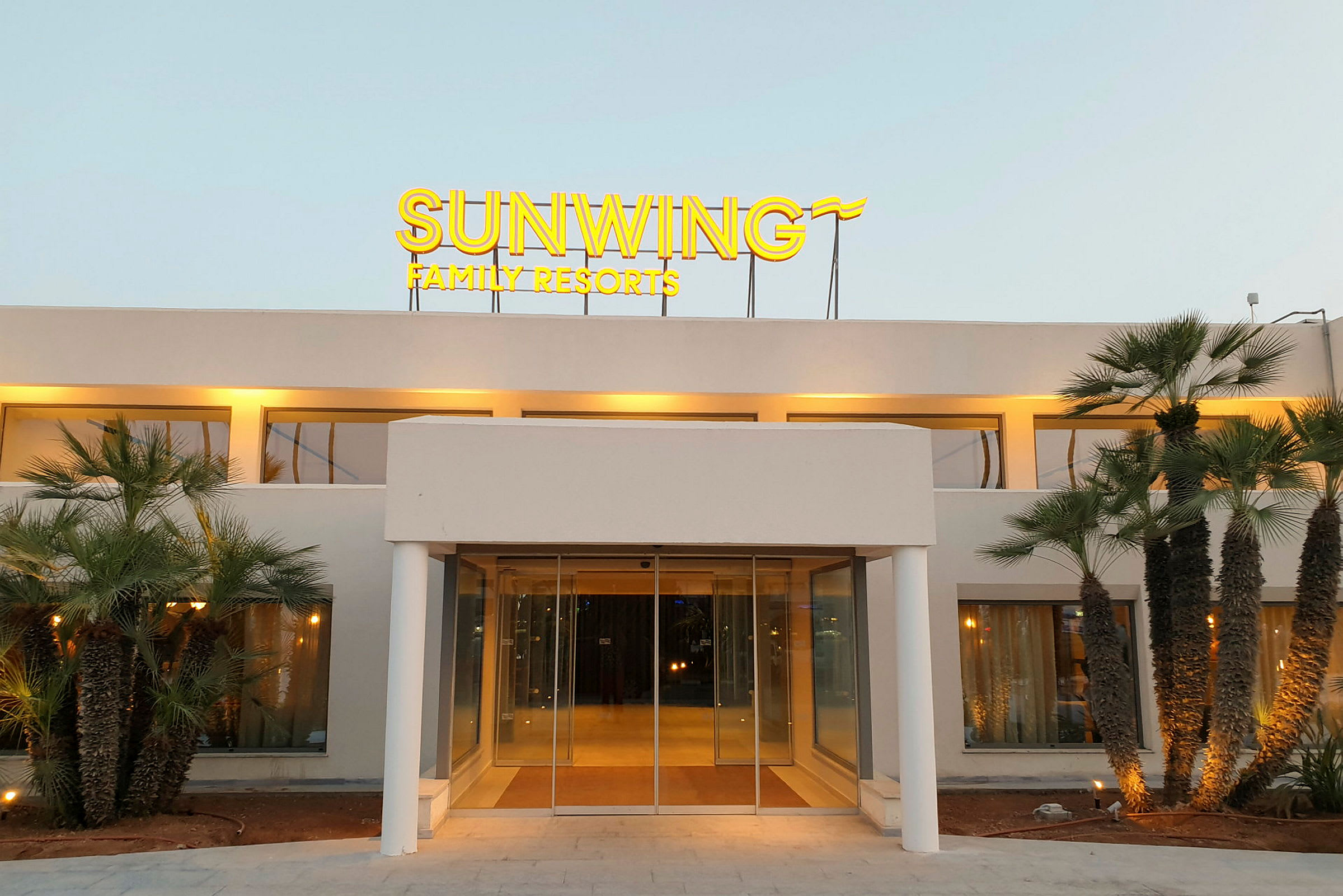 Forsiden af en bygning med et oplyst skilt, der siger "Sunwing Family Resorts", omgivet af palmer. (AI-genereret tekst)