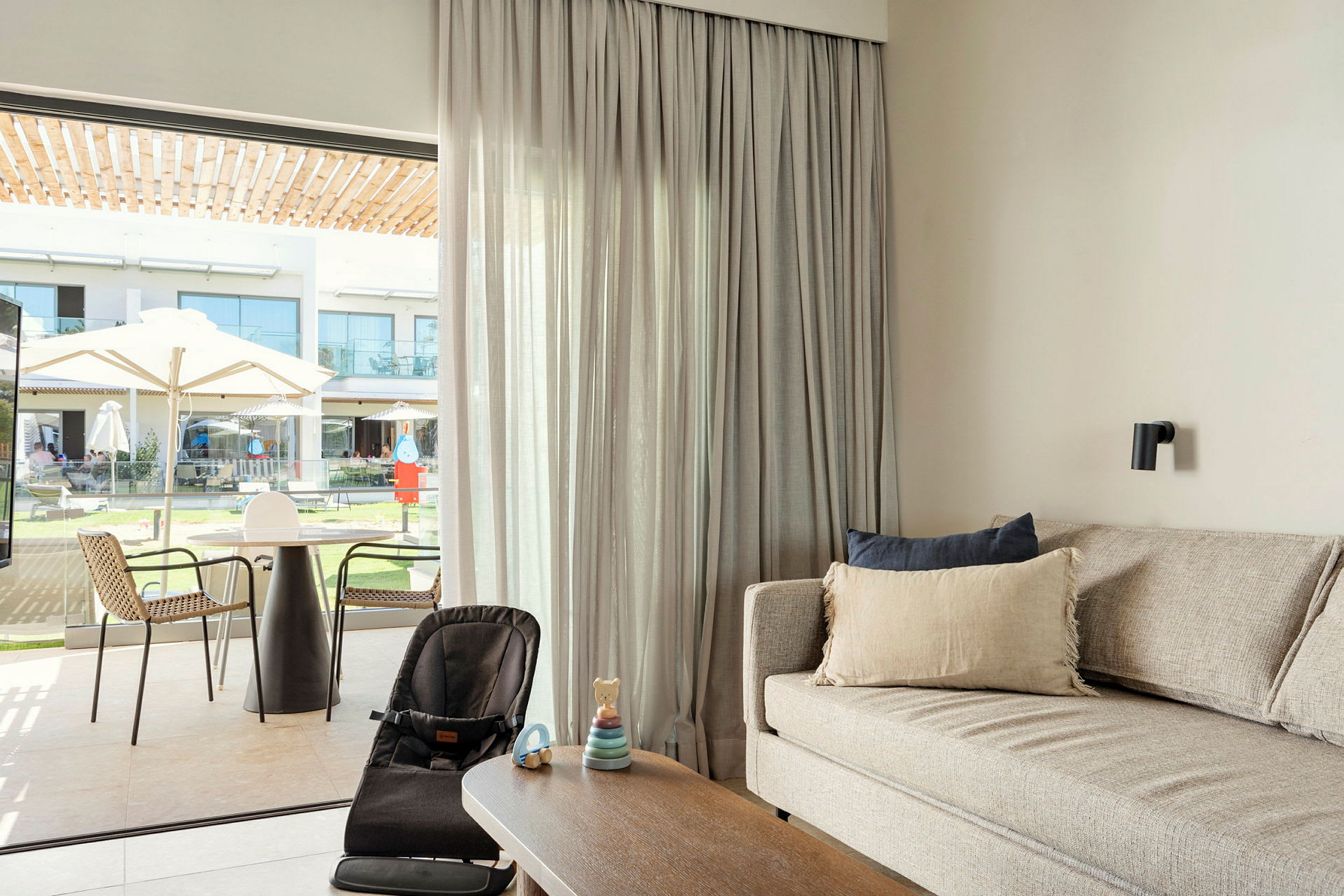 En moderne stue med en sofa, et sofabord og en babystol foran et stort vindue, der vender ud mod en udendørs terrasse og have. (AI-genereret tekst)