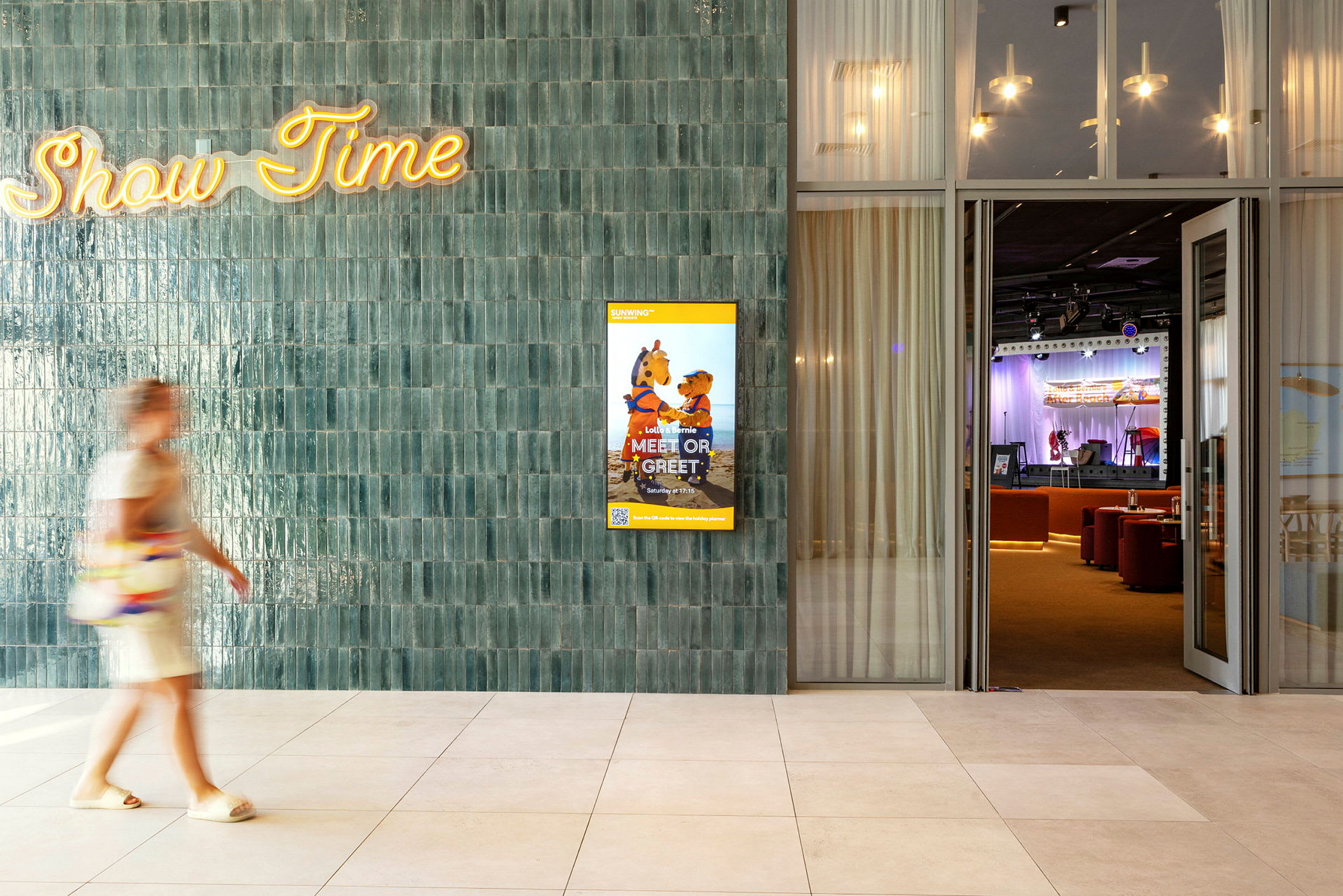 En person går forbi en væg med teksten "Show Time" og en plakat for et børneprogram. I baggrunden ses en scene med publikumsstole. (AI-genereret tekst)