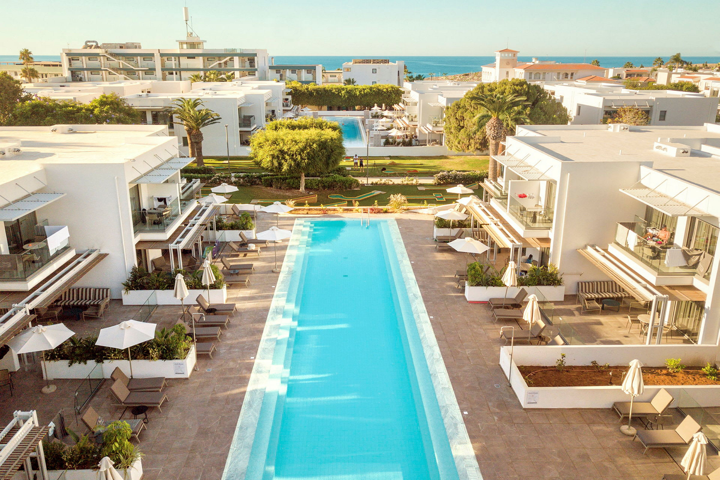 Et hotelområde med en lang swimmingpool mellem to hvide bygninger. I baggrunden ses frodige græsplæner og havet. (AI-genereret tekst)