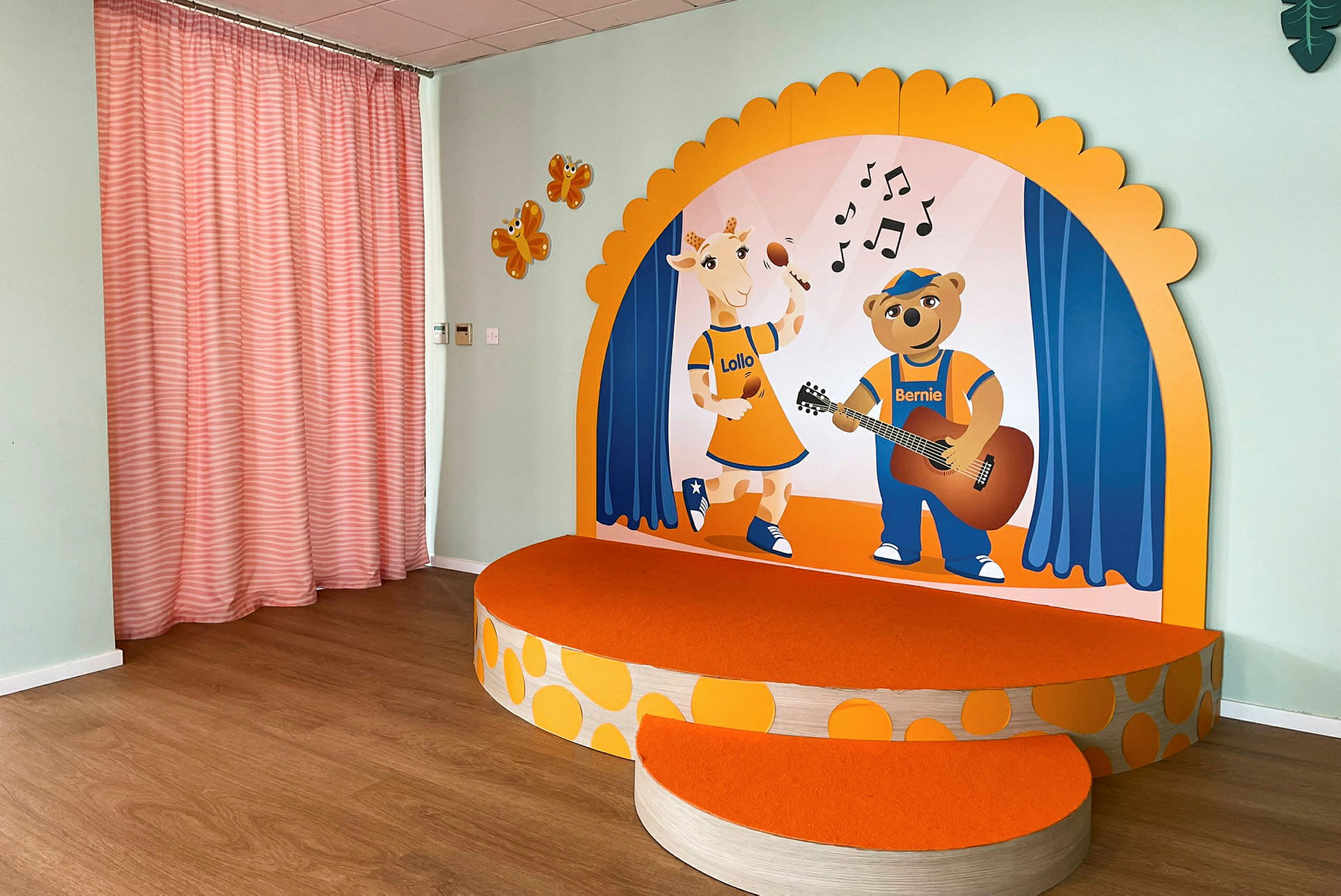 Et lille podie med orange overflade og illustrationer af en giraf og en bjørn på en baggrund med musikteatertema. På sidevæggen er der orange figurer og lyserøde gardiner. (AI-genereret tekst)