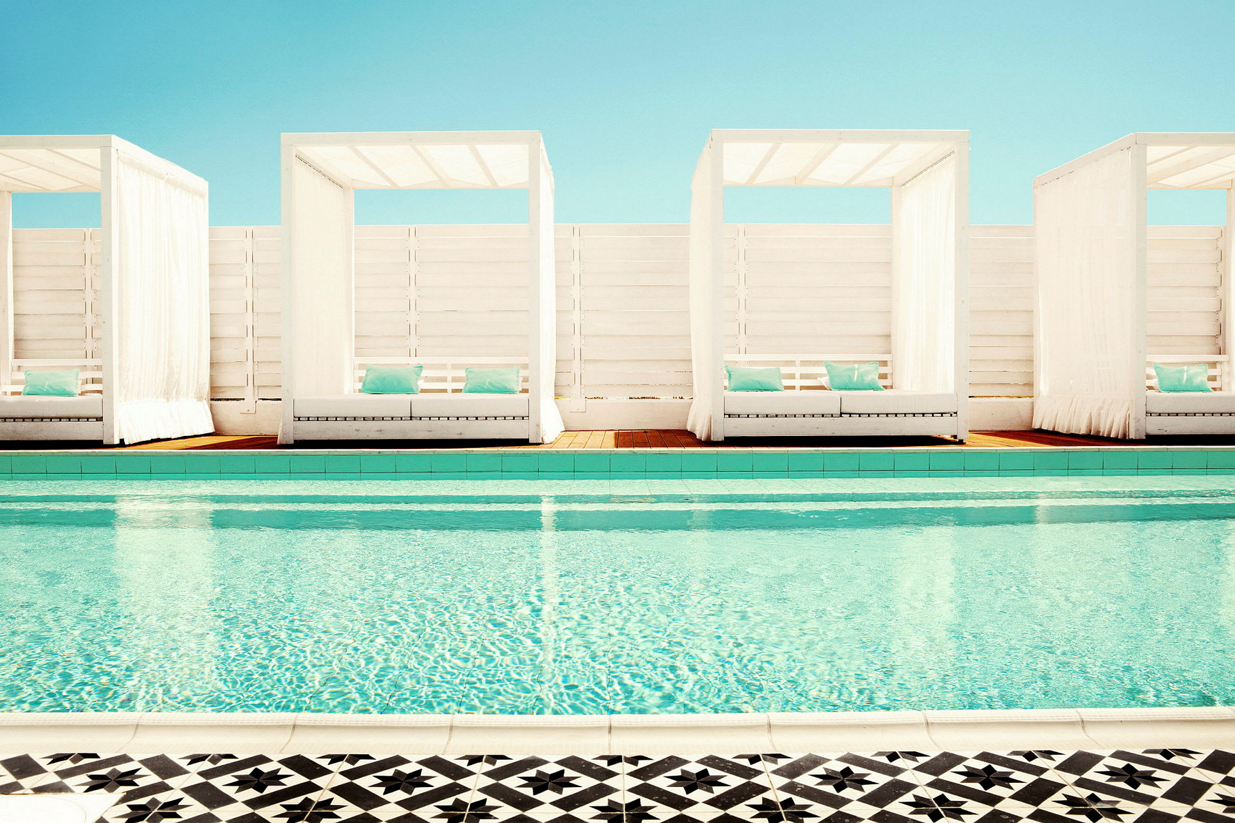 En pool med klart vand og tre cabanas med hvide stoffer og blå puder i baggrunden. (AI-genereret tekst)