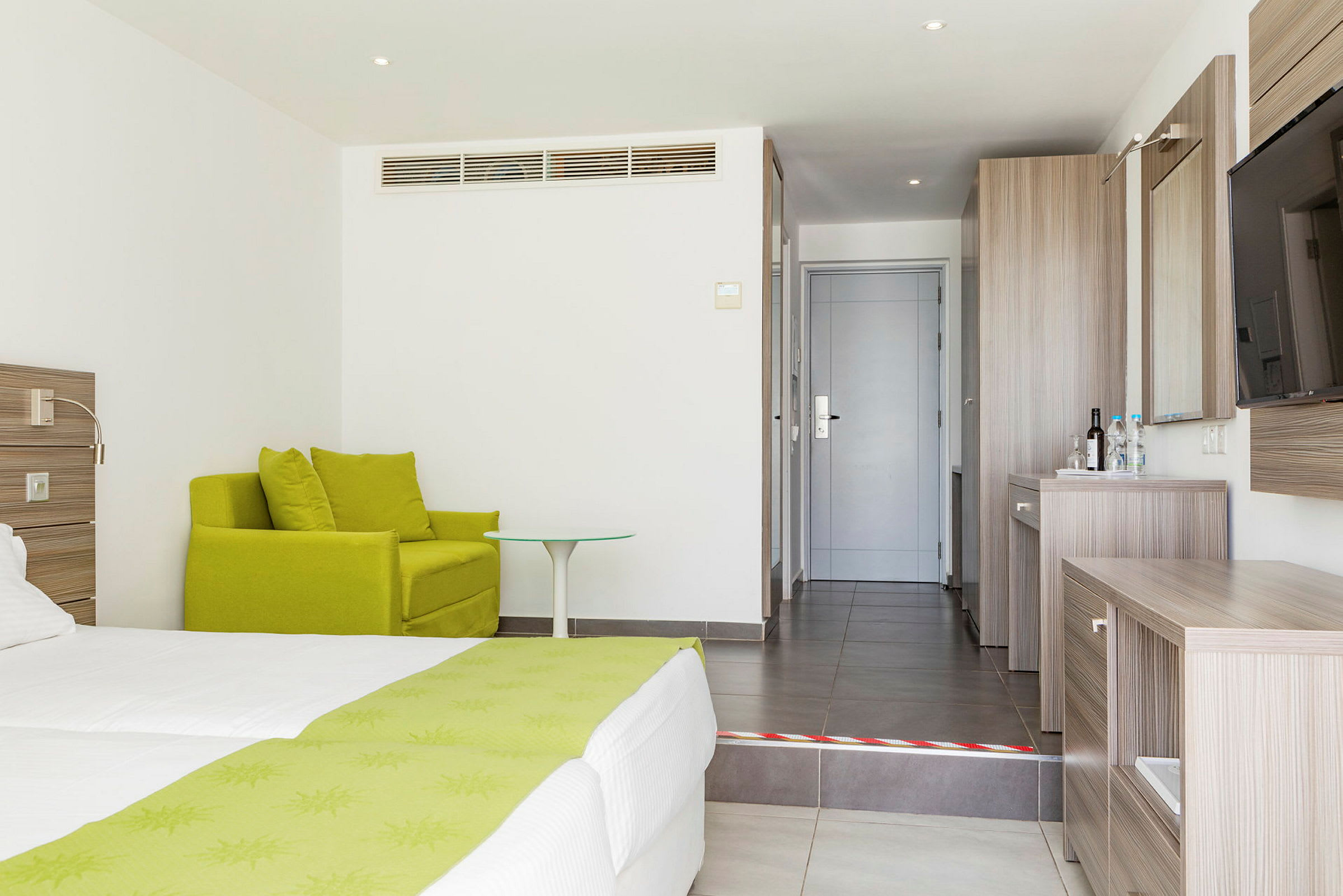 Hotelværelse med grøn lænestol, seng med grønne detaljer og træskrivebord ved indgangen. (AI-genereret tekst)