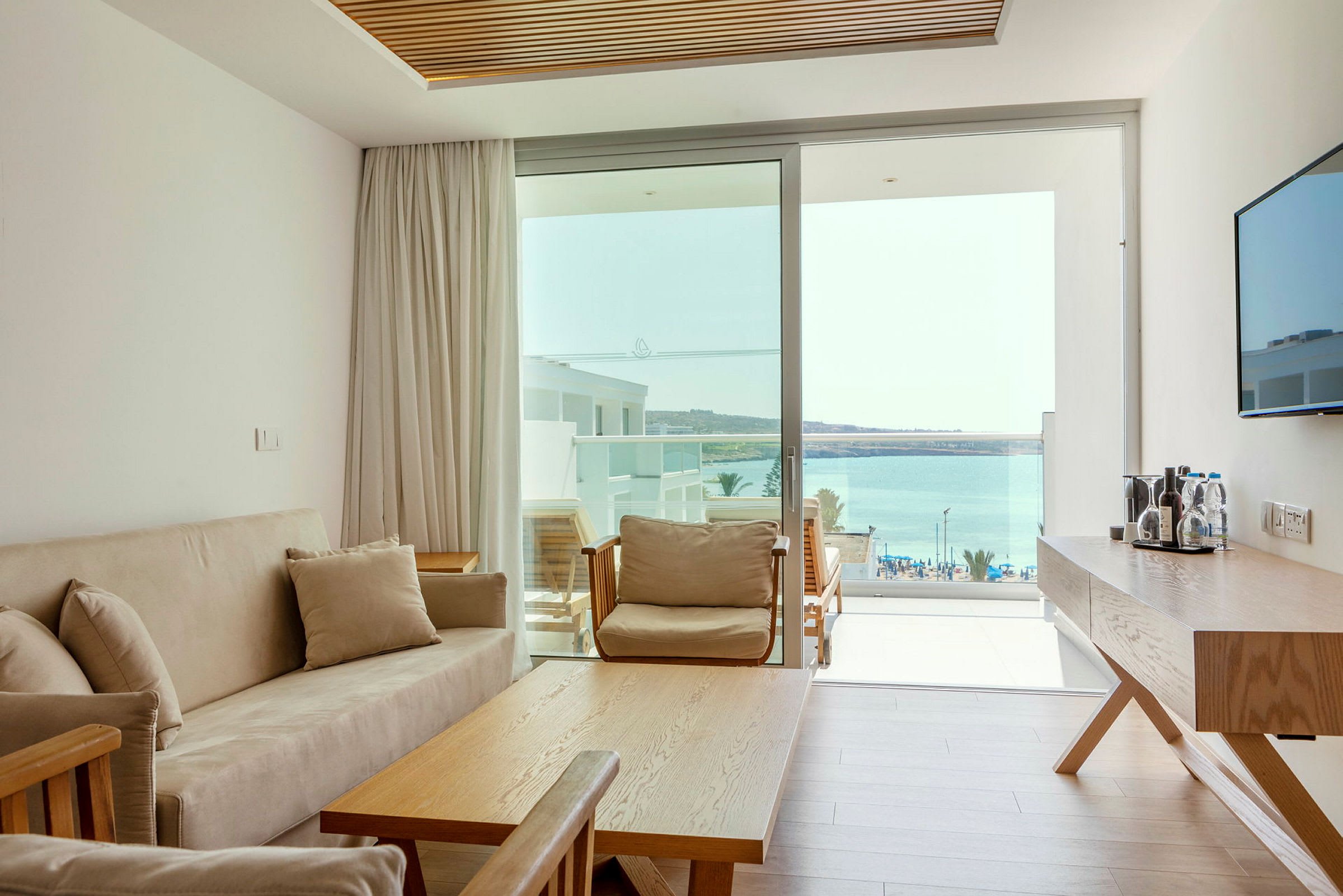 Et lyst hotelværelse med en sofa, bord og udsigt over havet gennem glasdøre. (AI-genereret tekst)