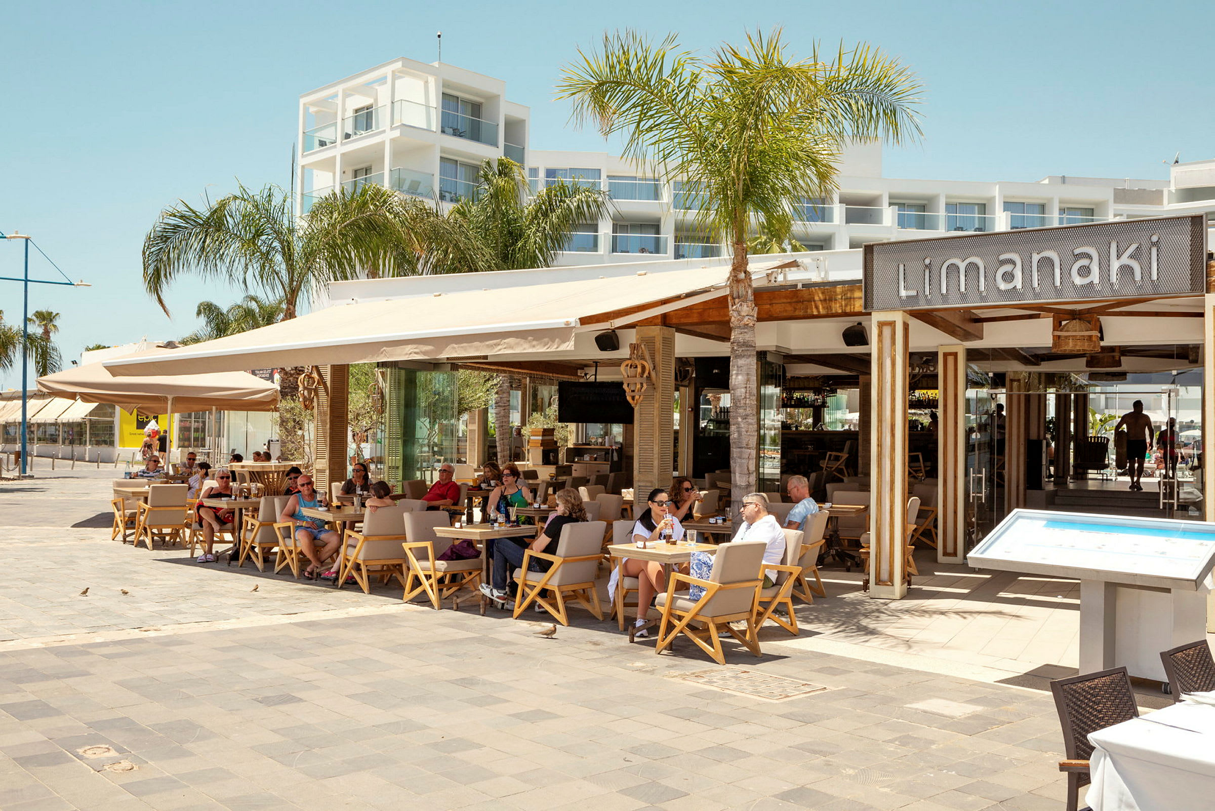 Restaurant Limanaki med siddende gæster, omgivet af palmer og moderne arkitektur. (AI-genereret tekst)