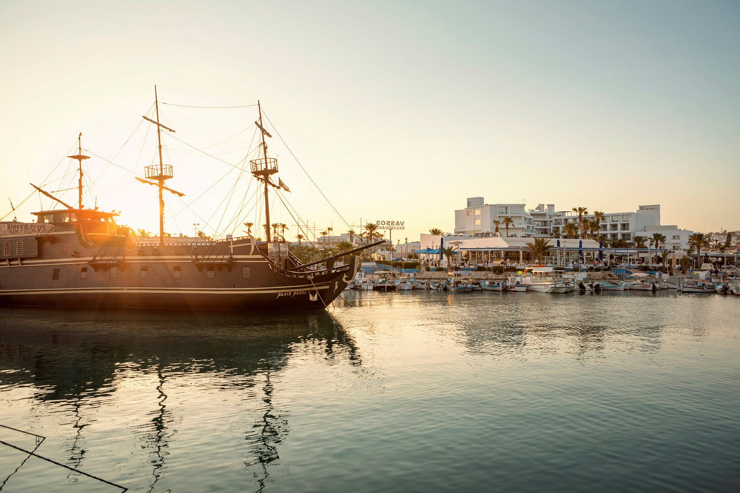 Et piratskib ligger i en havn med bygninger og både i baggrunden ved solnedgang. (AI-genereret tekst)