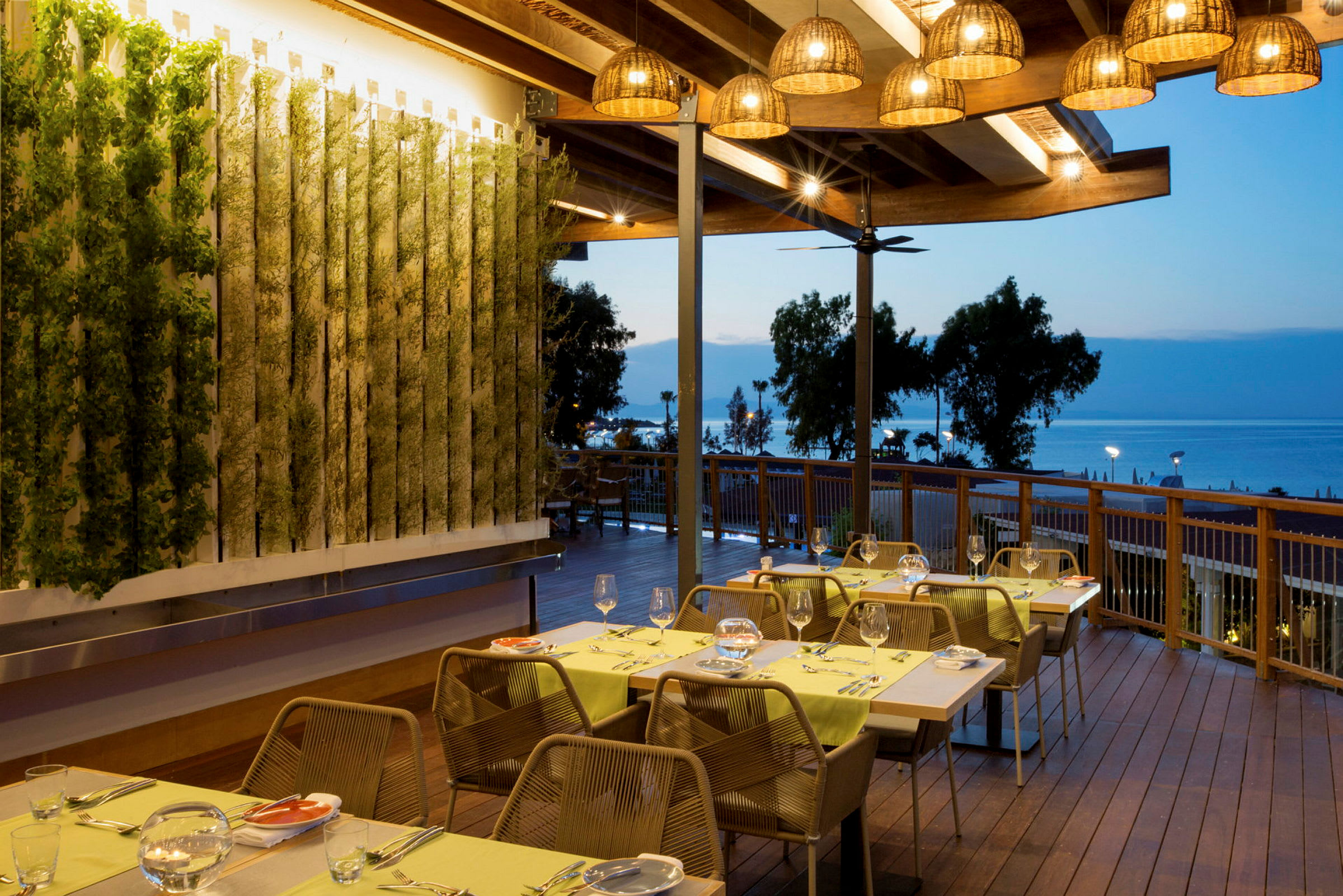 Restaurant med udendørsterrasse, borde dækket til middag og havudsigt i baggrunden. (AI-genereret tekst)