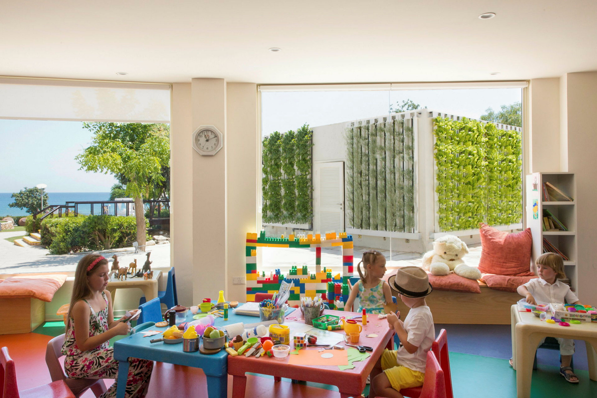Børn leger ved et bord med legetøj og aktiviteter indendørs, med udsigt til en have udenfor. (AI-genereret tekst)