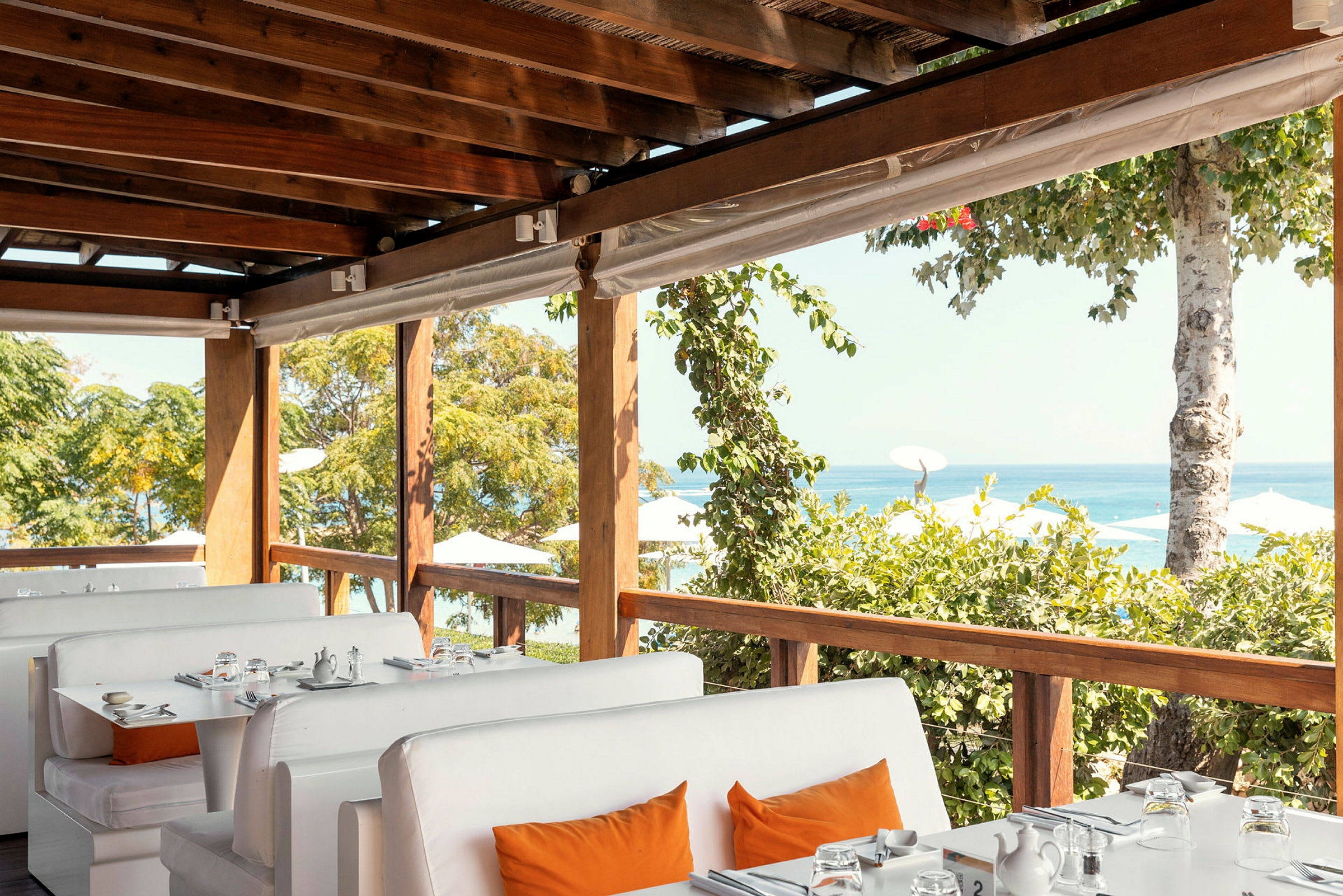 Billede af en restaurant med hvide sofaer og borde med udsigt over havet og bevoksning. (AI-genereret tekst)